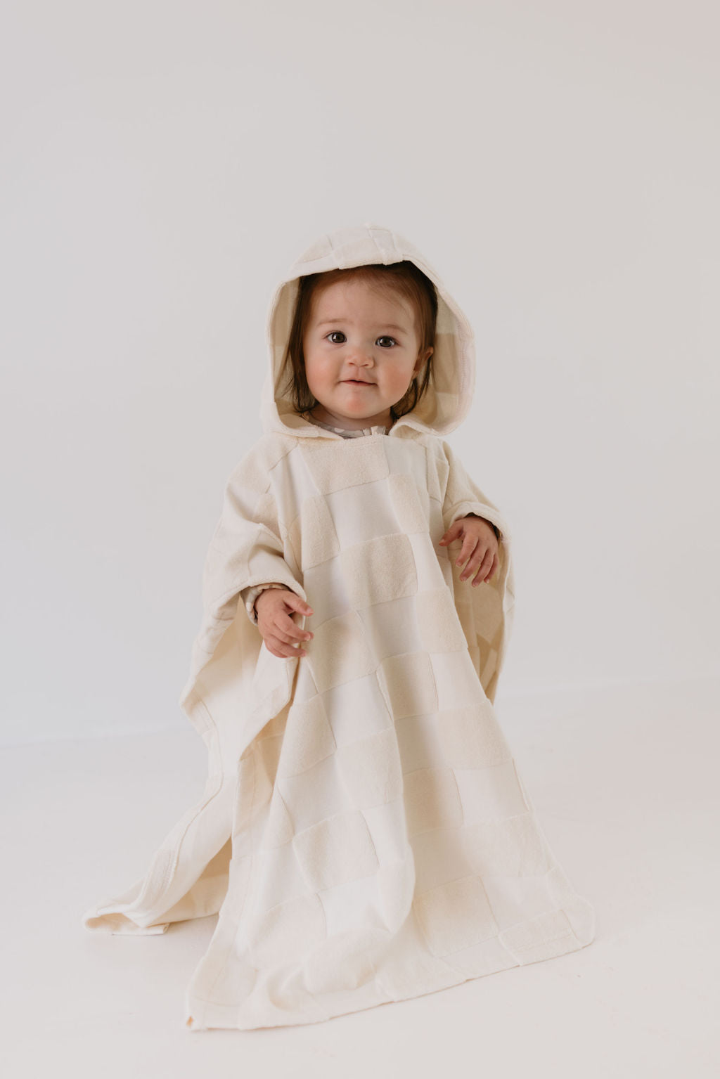 A baby wearing a cream checkerboard hooded towel by forever french baby stands against a plain white background, looking directly at the camera.