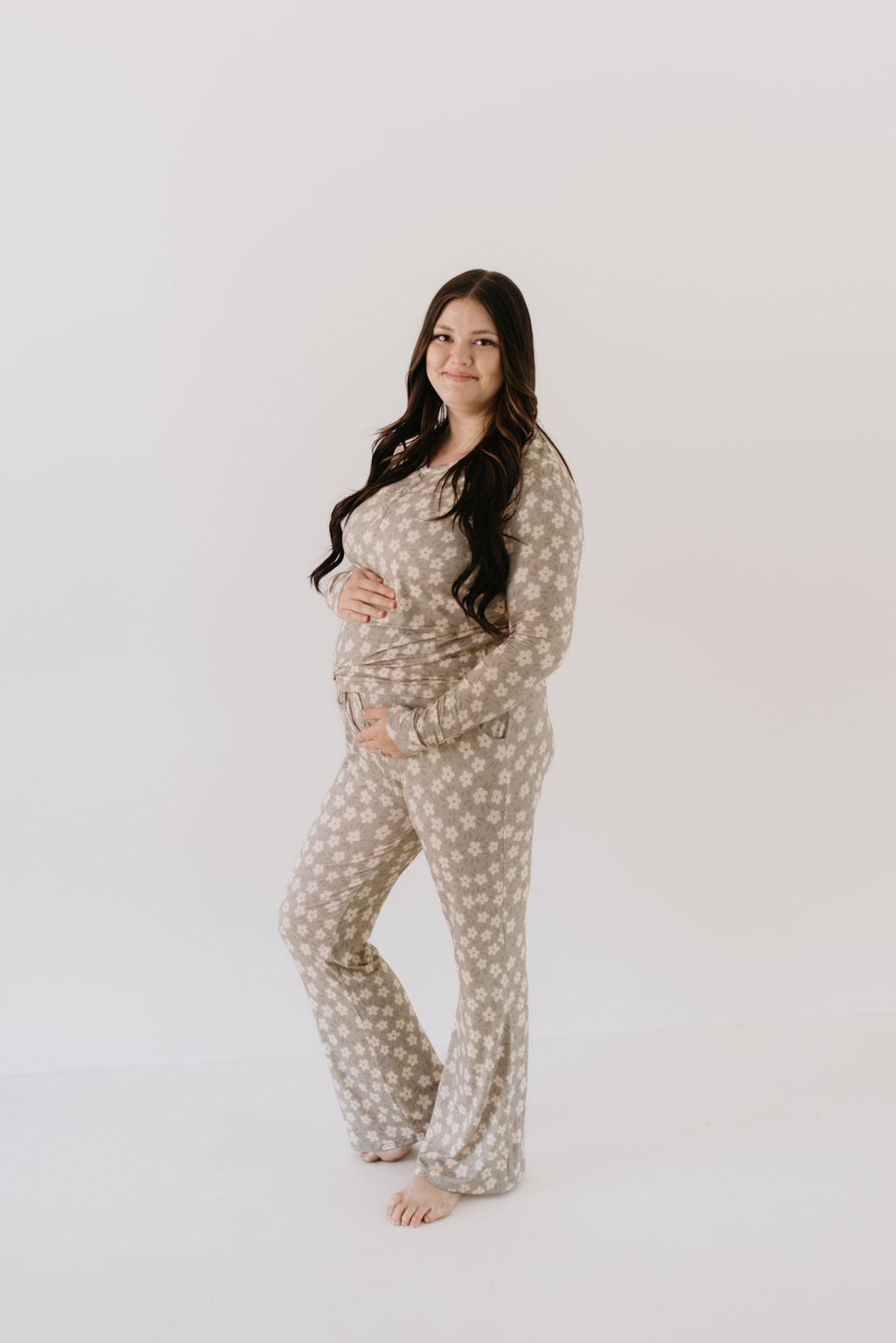 A pregnant woman with long hair stands barefoot, wearing a matching set of hypo-allergenic and breathable Women's Bamboo Pajamas in the French Gray Floral pattern by forever french baby. The fabric gently drapes her figure as she holds her belly and smiles against a plain white background.