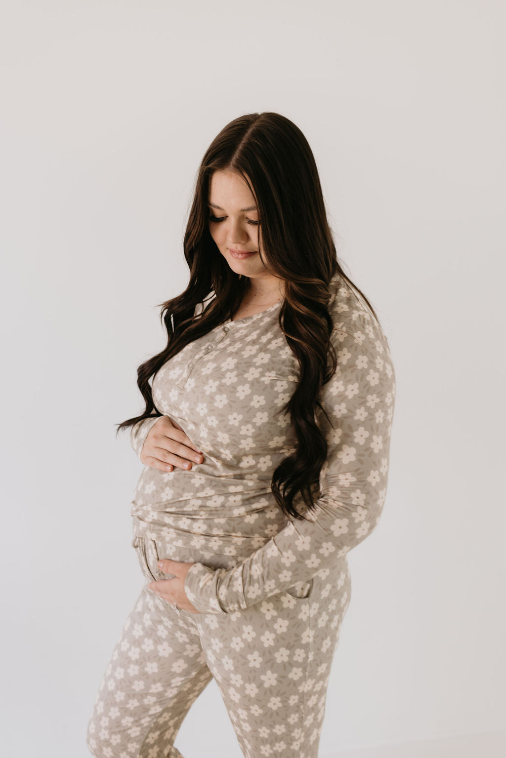 A woman with long, dark hair stands gracefully, admiring her baby bump and gently cradling it while dressed in *forever french baby's* Women's Bamboo Pajamas in the French Gray Floral pattern. She exudes comfort against the plain white backdrop, embracing the softness of the breathable fabric.