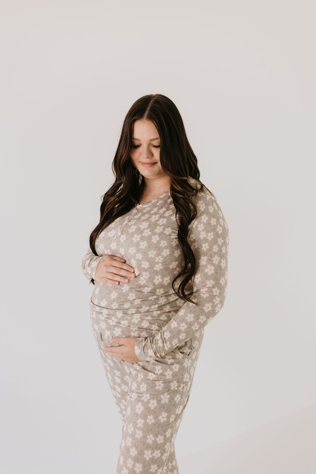 A pregnant woman with long dark hair gently touches her belly, dressed in a serene expression against a plain background. She wears the Women's Bamboo Pajamas in French Gray Floral by forever french baby, crafted from breathable fabric with a delicate white floral pattern.