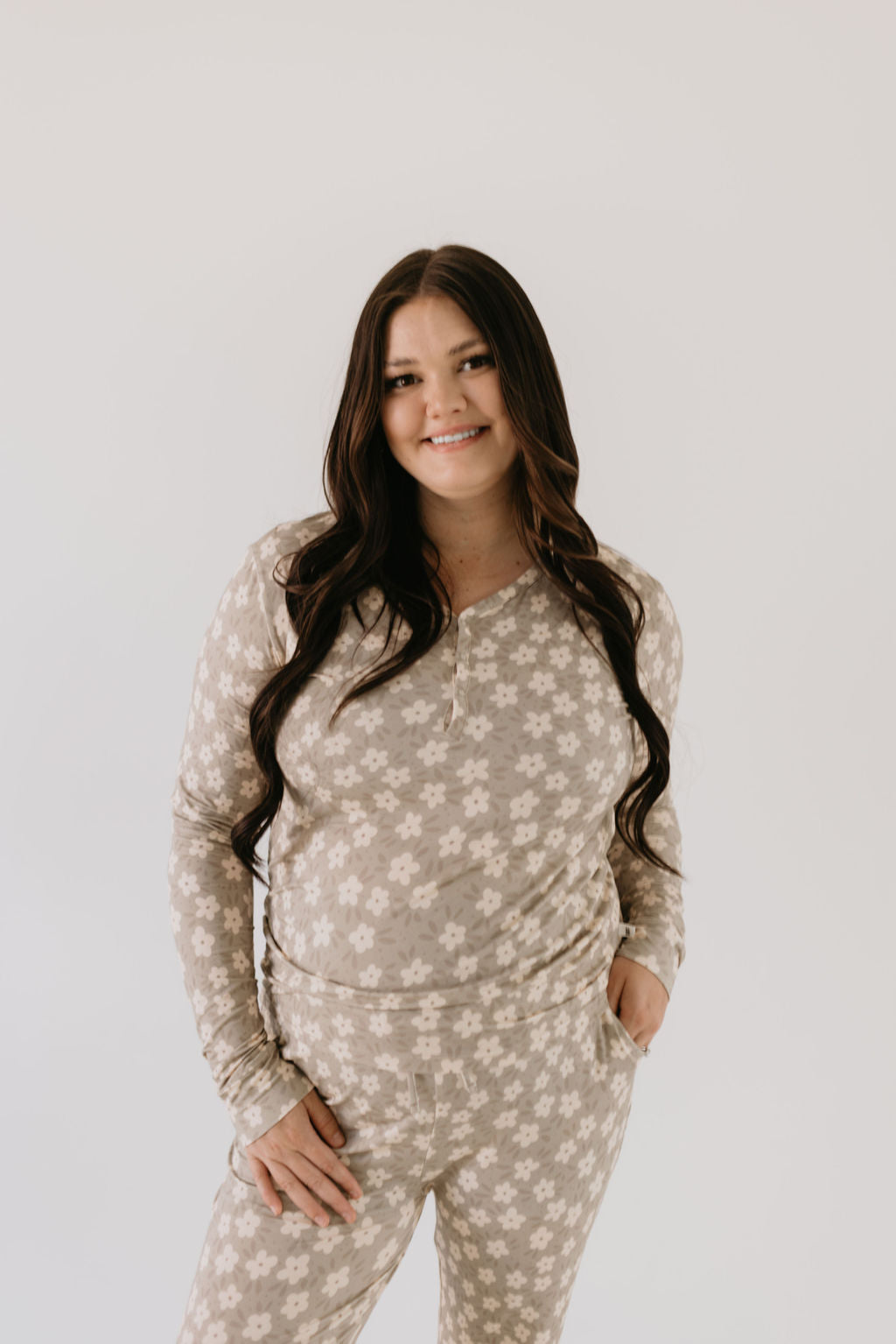 A woman with long dark hair stands against a plain background, wearing a matching outfit in breathable fabric from forever french baby. She has one hand in her pocket and is smiling slightly, showcasing the Women's Bamboo Pajamas in French Gray Floral.