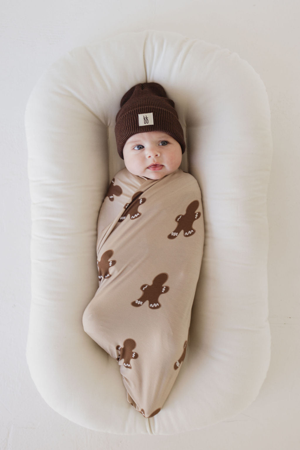 A baby is swaddled in the Bamboo Swaddle | Gingerbread by forever french baby, featuring charming brown gingerbread prints, and wears a soft brown beanie. Resting on a white cushion, the little one looks slightly to the side, wrapped in the comfort of hypo-allergenic materials.