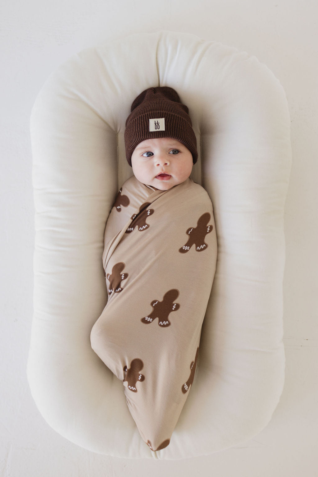 A baby lies wrapped in the hypo-allergenic Bamboo Swaddle | Gingerbread by forever french baby, donning a brown beanie and resting comfortably on a soft, white cushion.