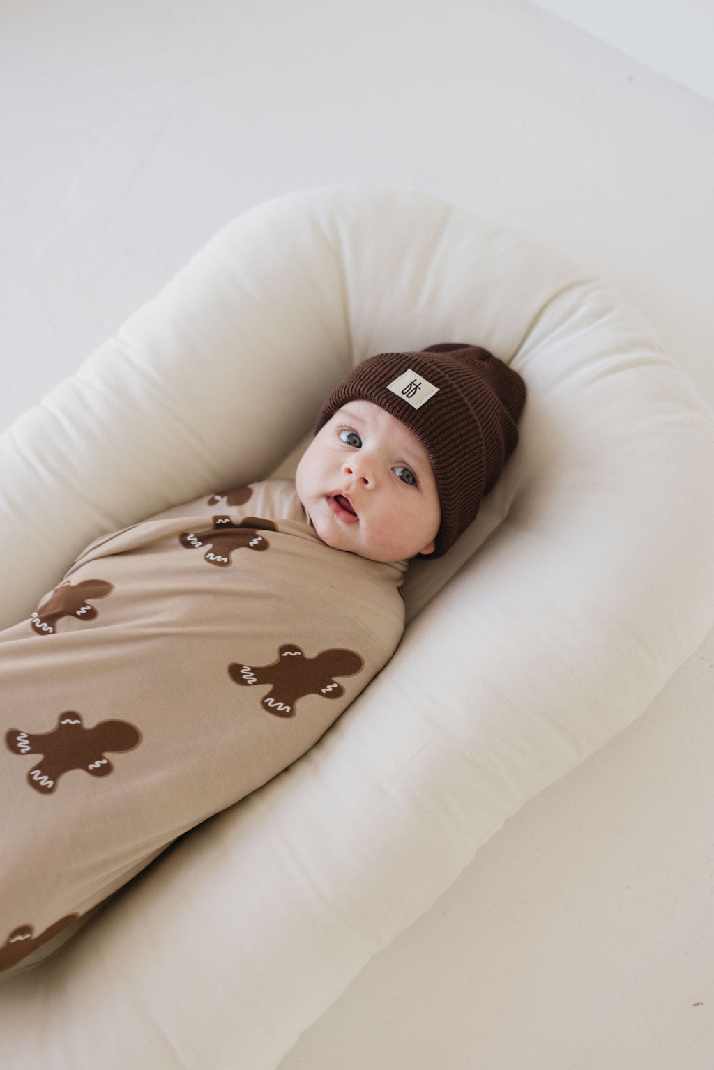 A baby wrapped in the Bamboo Swaddle | Gingerbread by forever french baby lies on a soft, white pillow. The hypo-allergenic fabric ensures comfort while the baby, wearing a brown beanie, looks up with wide eyes.