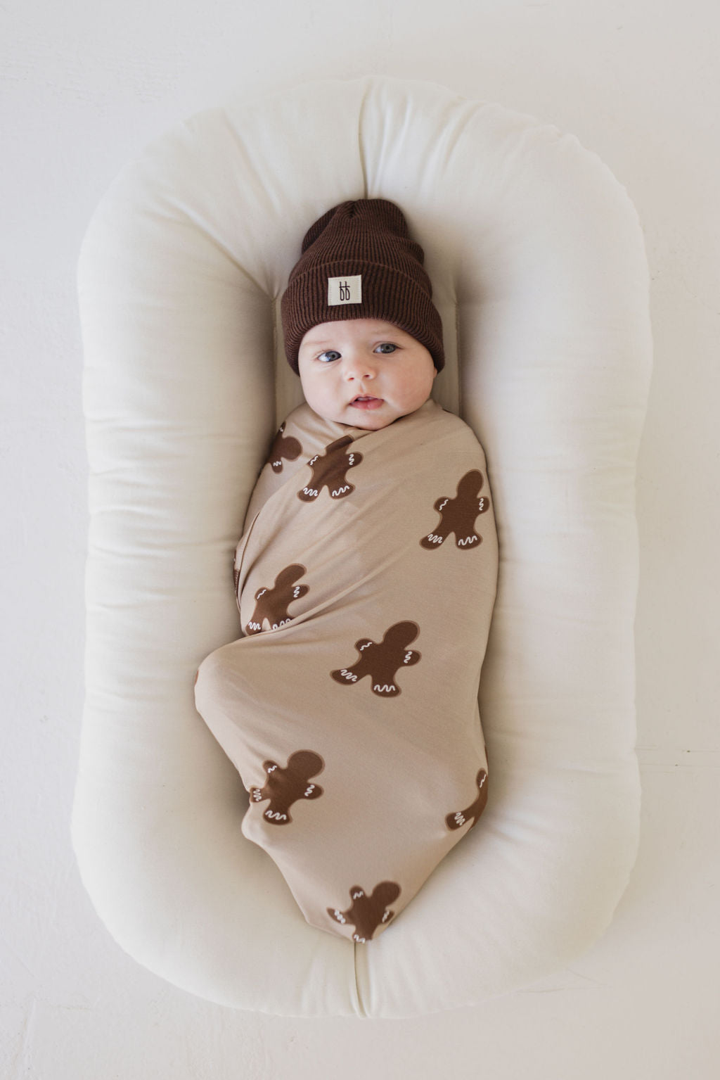 A baby is gently swaddled in a Forever French Baby Bamboo Swaddle in the Gingerbread design, featuring hypo-allergenic materials and brown teddy bear prints. The baby lies peacefully in a white cushioned nest, wearing a cozy brown knit hat with a calm expression against a light, soft background.