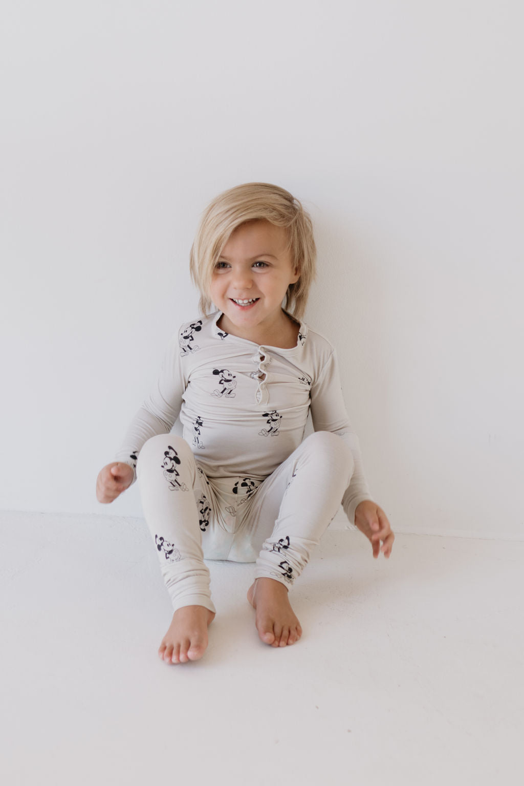 A young child with blonde hair is seated on the floor against a white background, dressed in the light-colored, playful print Bamboo Two Piece Pajamas - All Aboard! by forever french baby. The outfit's hypo-allergenic bamboo fabric ensures comfort as the child smiles and glances slightly to the side.