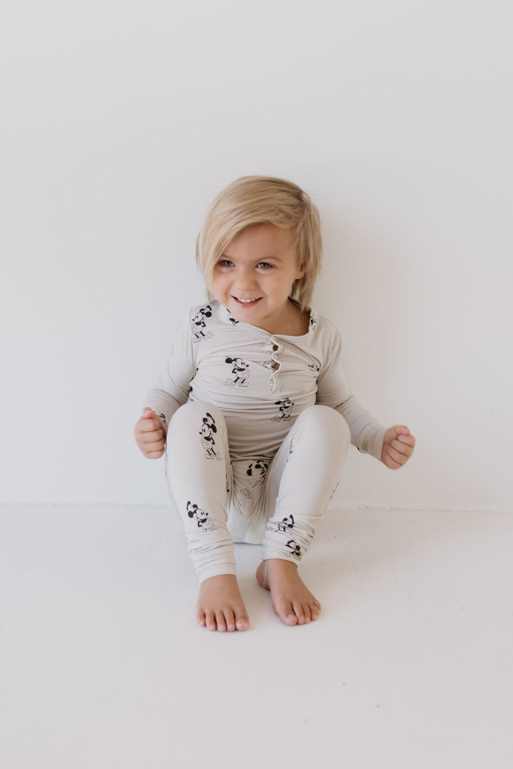 A young child with blonde hair sits against a white wall, smiling in their light-colored Bamboo Two Piece Pajamas named "All Aboard!" by forever french baby. The playful pattern perfectly complements the room's bright, minimalistic atmosphere.