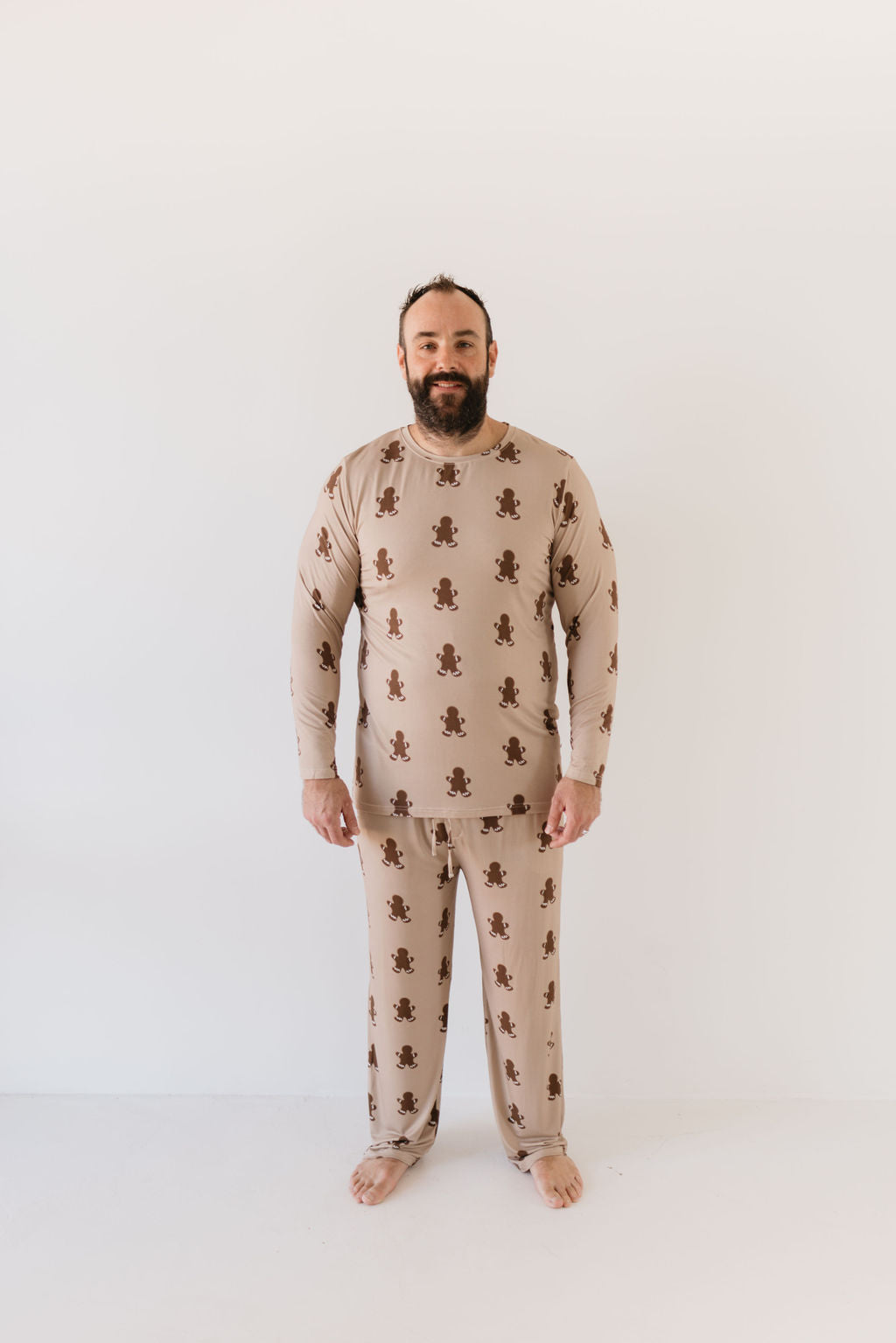 A bearded man smiles, standing barefoot in the Men's Bamboo Pajamas | Gingerbread by forever french baby. The hypo-allergenic clothing, made from breathable fabric, stands out against the plain white background.