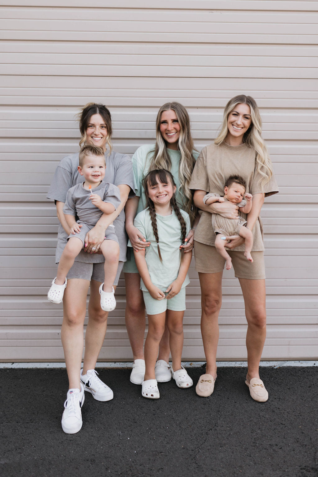 Four women and three children stand in front of a beige garage door, all dressed in neutral-toned casual outfits. Two children are carried by two women sporting the Vintage Washed Espresso Children's Short Set from Forever French Baby, while one young girl with braided hair, smiling in her children's summer outfit, stands in the center.