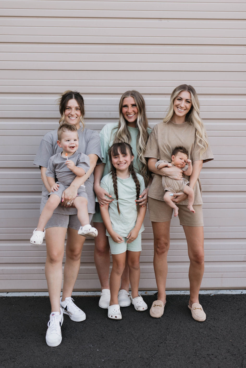 A group of three women and three children stand in front of a beige garage door, smiling. Two women are holding babies, while the third child stands in the middle with braided hair. They are all dressed in casual, matching neutral-colored outfits featuring the Women's Short Set | Vintage Washed Steel by forever french baby for a cozy yet stylish look.