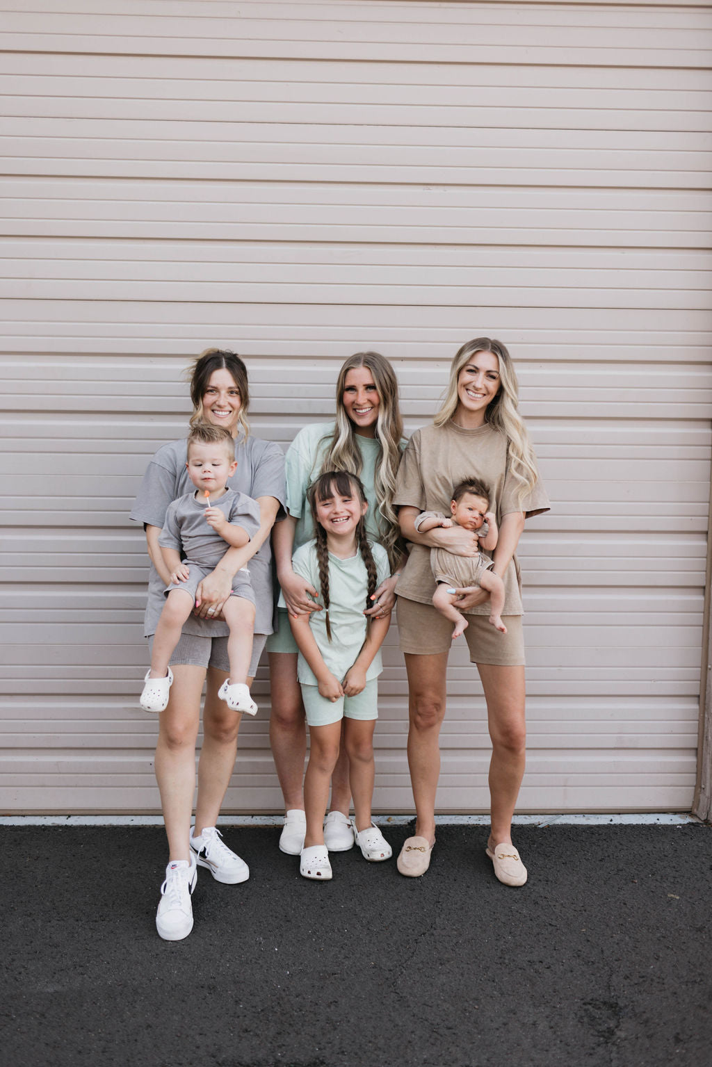 Three women and three children stand together in front of a beige garage door. Each woman holds a young child, and a girl with two braids stands in the middle. They are all casually dressed in neutral-toned outfits, including a Women's Short Set in Vintage Washed Spearmint and charming attire from forever french baby, smiling at the camera.