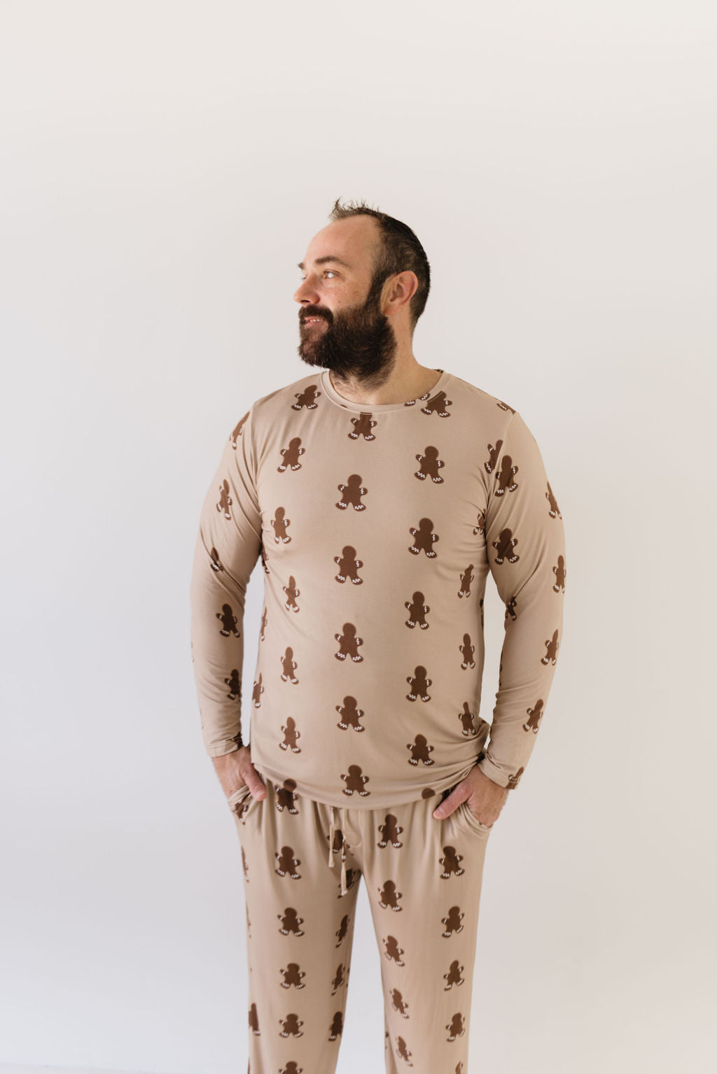 A man with a beard poses casually in the Men's Bamboo Pajamas | Gingerbread from forever french baby. Crafted from breathable fabric, these beige pajamas are adorned with gingerbread men, offering ultimate comfort against the plain white background—ideal for relaxation.