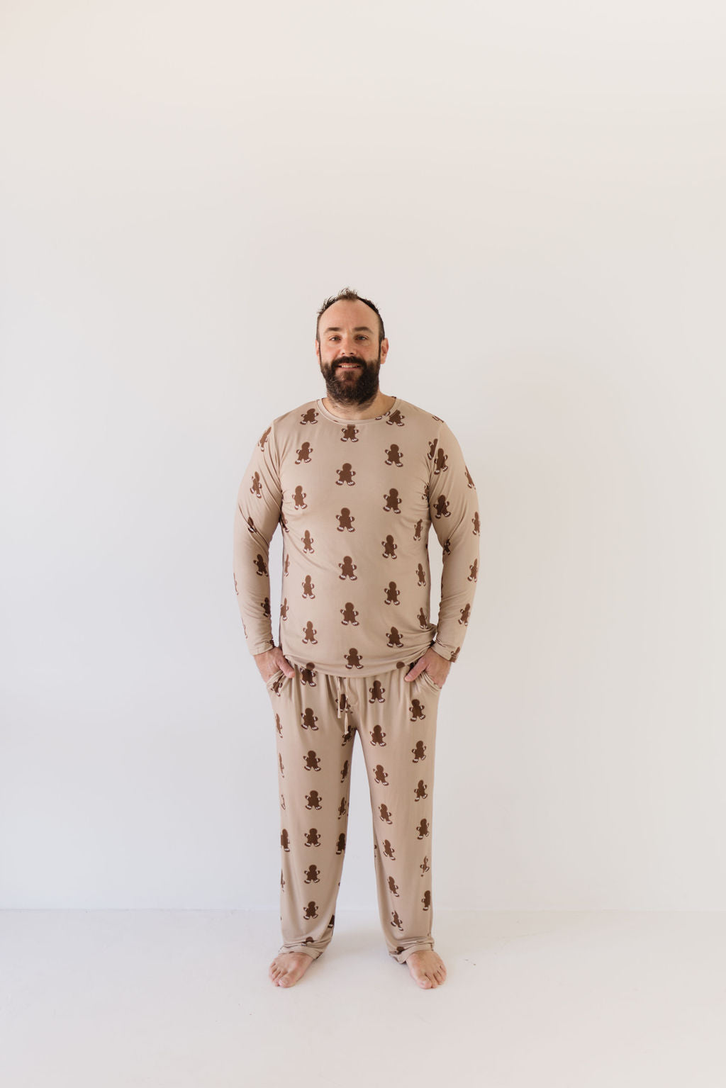 A bearded man stands in a neutral pose against a plain background, wearing the Men's Bamboo Pajamas in Gingerbread from forever french baby. The pajamas are long-sleeved with matching beige fabric adorned with small teddy bear patterns, made from breathable material. He is barefoot and looking directly at the camera.