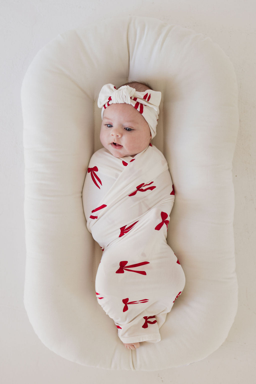 A baby is gently wrapped in the "Bamboo Swaddle | Ribbons & Bows" by forever french baby, nestled comfortably in a soft, cushioned bassinet. The hypo-allergenic fabric of the breathable bamboo blanket keeps the little one content and cozy while wearing a coordinating headband adorned with red bow patterns.