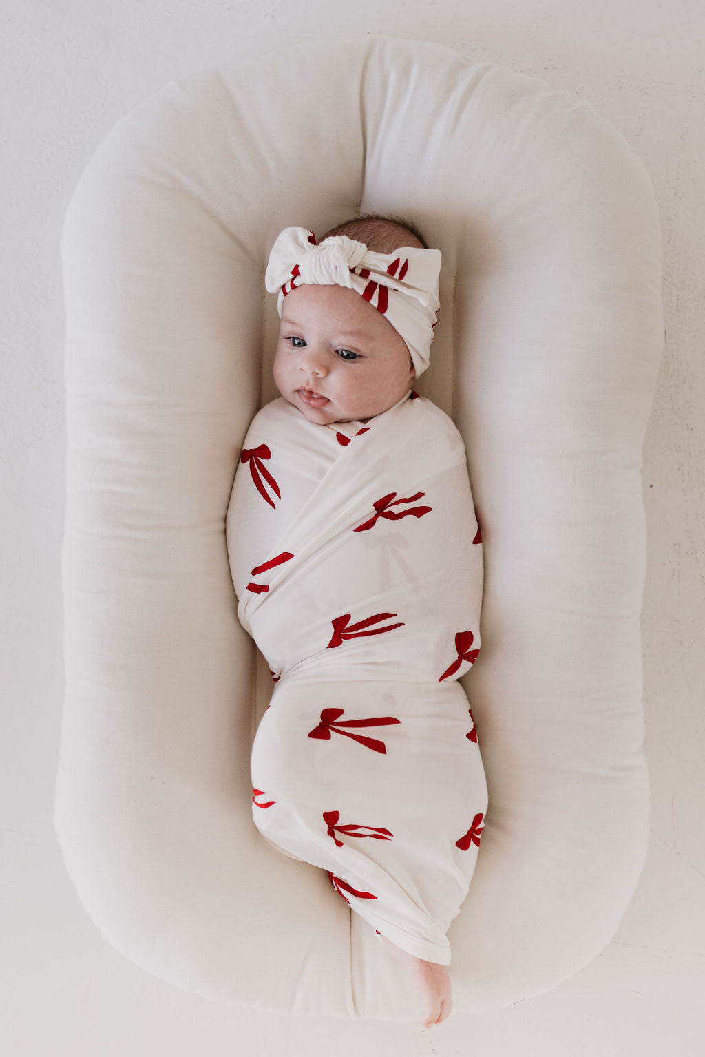 A baby snuggles in a cozy, hypoallergenic outfit featuring the "Ribbons & Bows" design by forever french baby, resting comfortably on a soft cushioned pad. The Bamboo Swaddle and matching headband provide breathability, enhancing the peaceful and serene atmosphere.