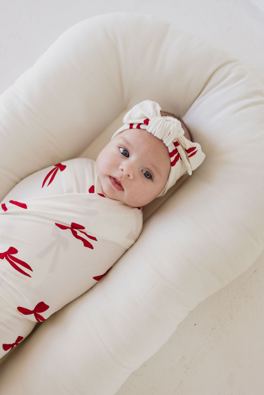 The baby is snugly wrapped in the Bamboo Swaddle | Ribbons & Bows from forever french baby, featuring red bow patterns, and lays comfortably in a soft, cushioned nest. A matching headband with a bow adorns the baby's head as they gaze up with a calm expression. The light, neutral background enhances the hypo-allergenic vibe.