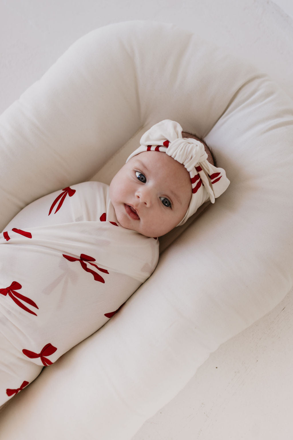 A baby wrapped in the "Bamboo Swaddle | Ribbons & Bows" from forever french baby, featuring breathable bamboo fabric adorned with red bow patterns, is resting on a soft, white cushion. The hypo-allergenic material ensures comfort and the baby is accessorized with a matching headband showcasing red stripes. The background is a light, neutral color.