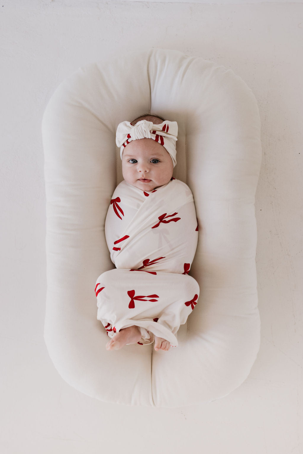 A baby is peacefully wrapped in a breathable, hypo-allergenic Bamboo Swaddle | Ribbons & Bows from forever french baby, resting on a white, cushioned surface. The baby wears a matching headband and gazes upward with a calm expression.