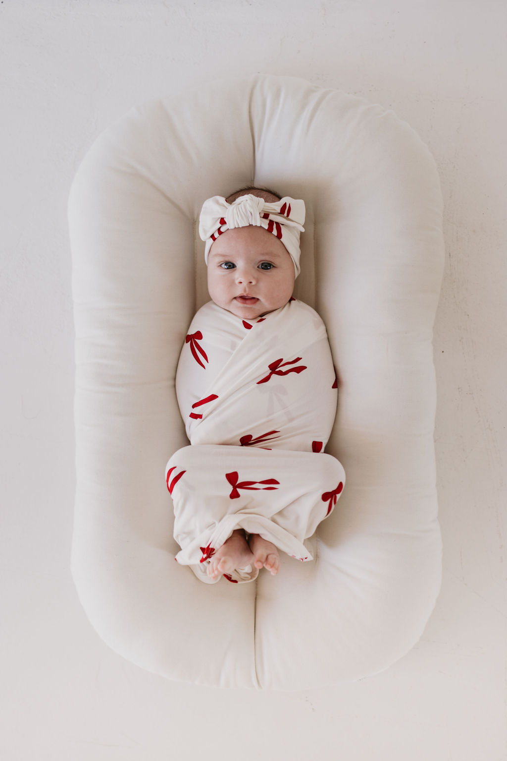 A baby wrapped in the Bamboo Swaddle | Ribbons & Bows by forever french baby lies on a white cushioned surface. The child is wearing a matching headband and looking upwards in a minimalistic and bright setting.