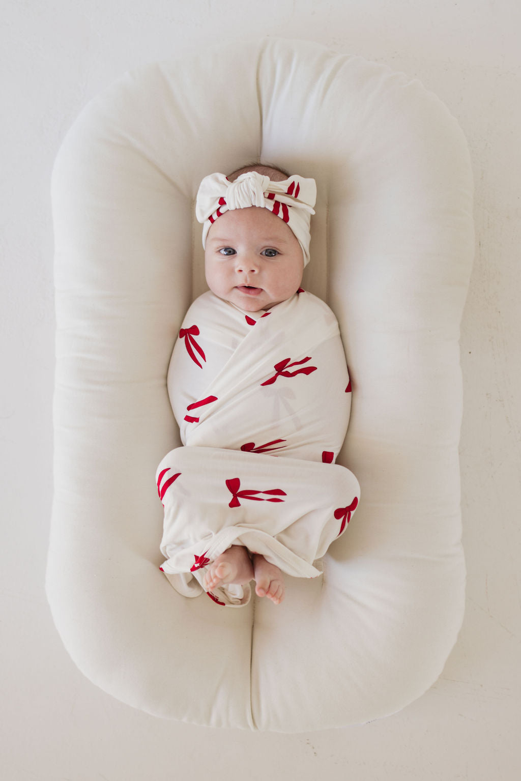 A baby rests on a plush white cushion, gently wrapped in a white blanket decorated with charming bow patterns. The infant wears the forever french baby Bamboo Head Wrap | Ribbons & Bows, featuring an adjustable head wrap with a matching bow, gazing up at the camera against a simple white backdrop.