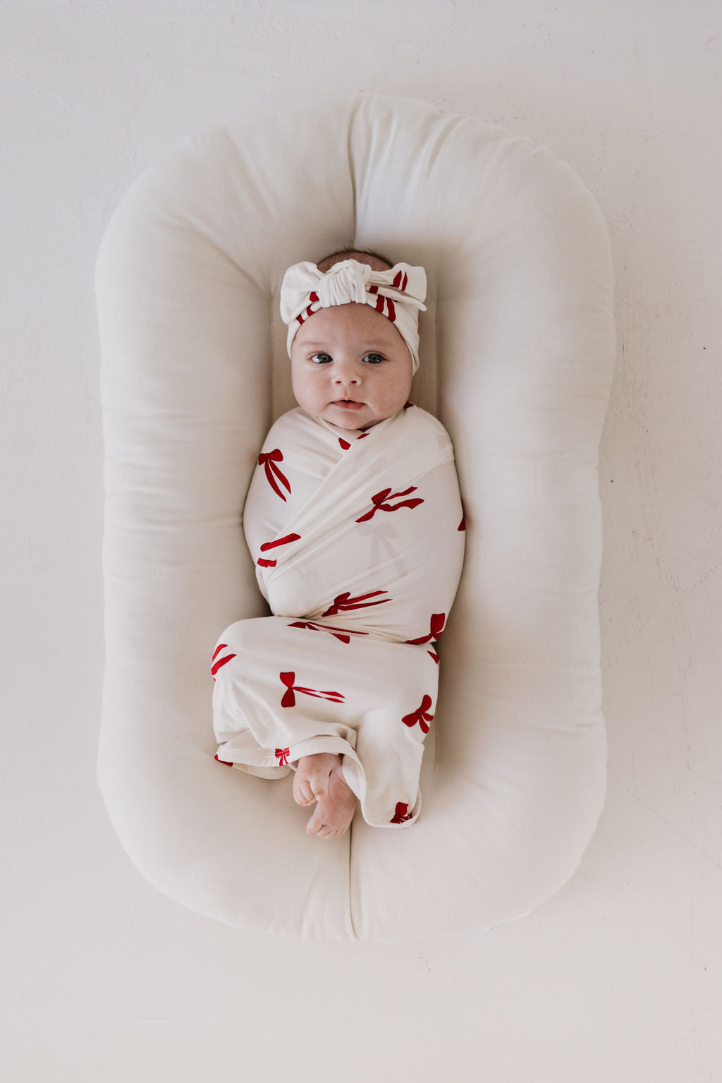 A baby lies on a plush white cushion wrapped in a swaddle featuring red bow patterns. The ensemble is beautifully complemented by the forever french baby Bamboo Head Wrap | Ribbons & Bows, perfectly matching the serene expression as they gaze upward.