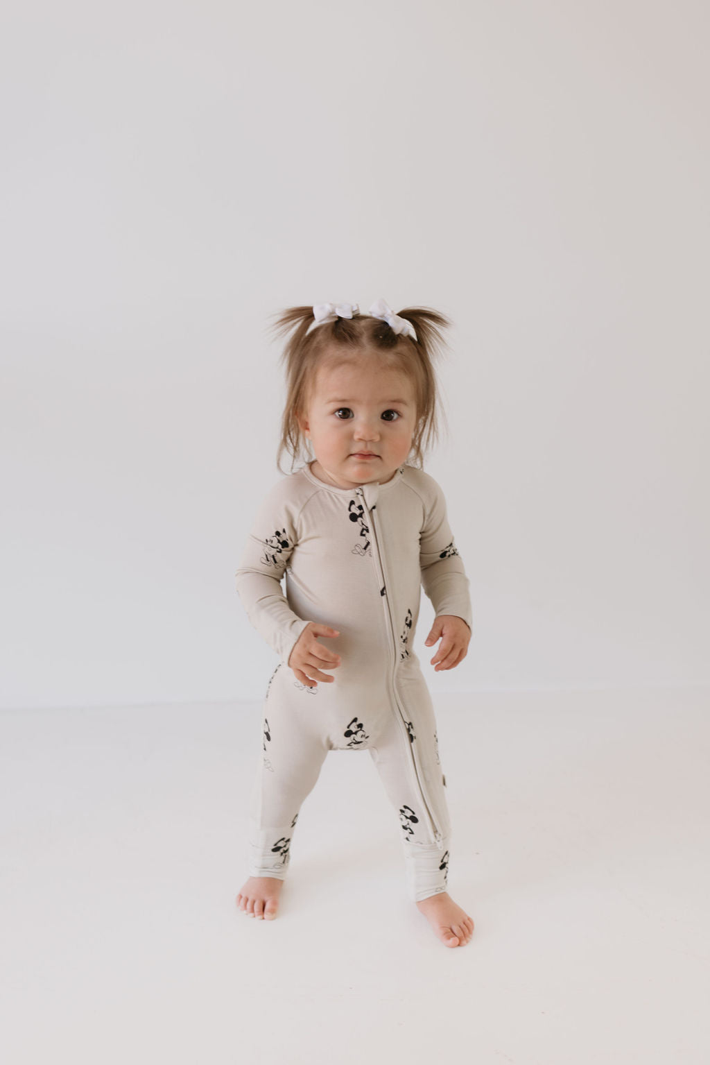 A toddler with brown hair styled in small pigtails is standing against a plain white background, dressed in breathable Bamboo Zip Pajamas from forever french baby, featuring black and white animal prints on a beige base.