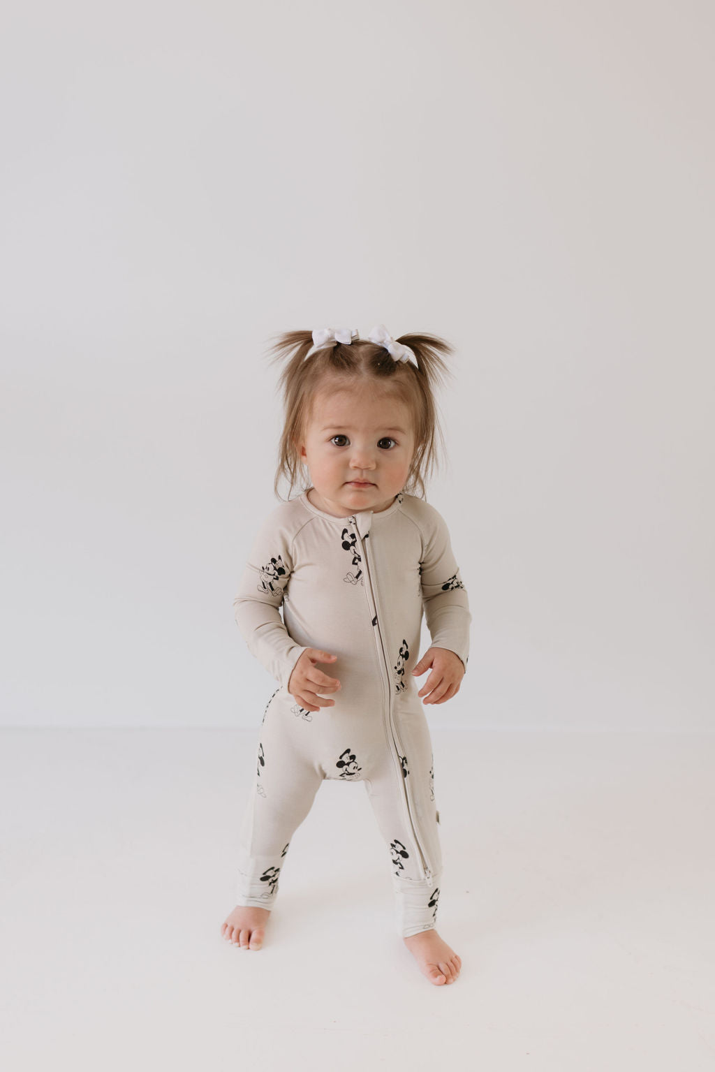 A toddler with two small ponytails wears Bamboo Zip Pajamas from the brand forever french baby, featuring a panda print. She stands against a plain white background and looks directly at the camera with a neutral expression.