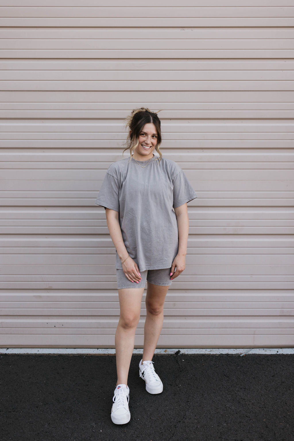 A woman stands in front of a beige rolling garage door, wearing the forever french baby Women's Short Set | Vintage Washed Steel. Her loose-fitting gray T-shirt and ribbed biker shorts complement her outfit perfectly. Her hair is tied up, and she completes her look with white athletic shoes and a relaxed, slightly smiling expression with one hand resting on her hip.