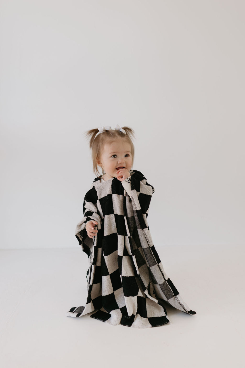 A toddler with small ponytails sits on the floor, wrapped in an oversized Hooded Towel in Black Checkerboard by forever french baby. The child is looking forward with a curious expression while holding the towel close. The background is plain white.