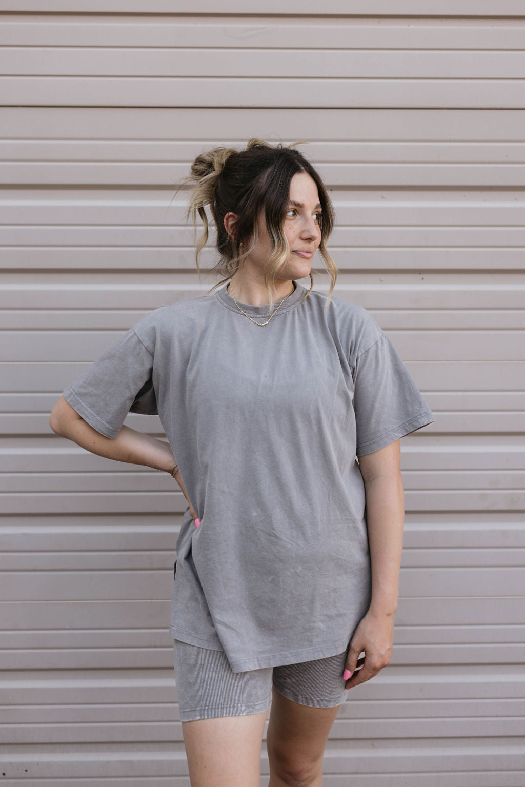 A person with light hair stands in front of a white corrugated metal wall. They are wearing the Women's Short Set | Vintage Washed Steel from forever french baby, featuring a grey t-shirt and matching ribbed biker shorts. One hand is on their hip, and they are looking to the side with a slight smile.