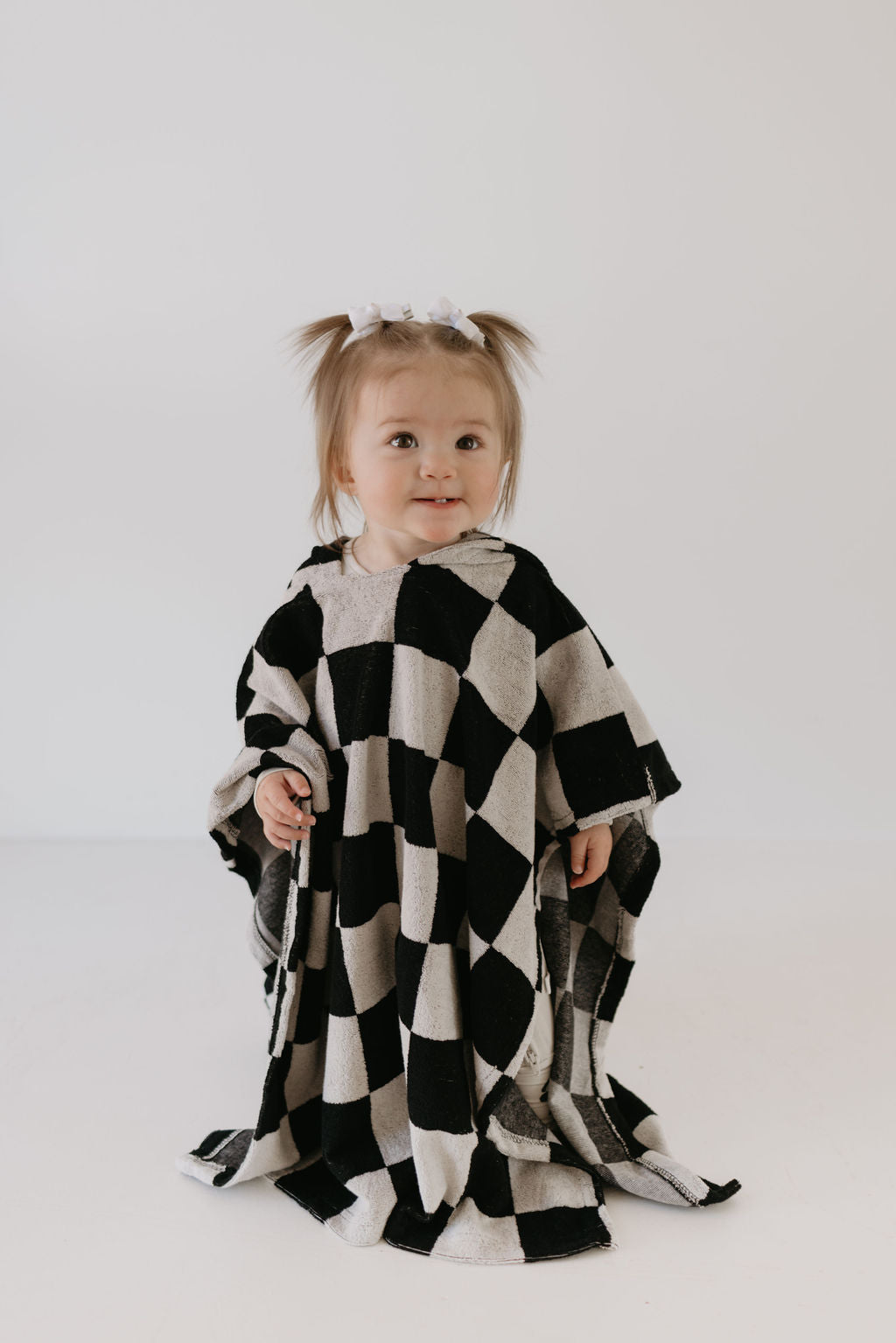 A toddler with pigtails is wrapped in the Hooded Towel | Black Checkerboard from forever french baby. She stands against a plain white background, looking towards the camera.