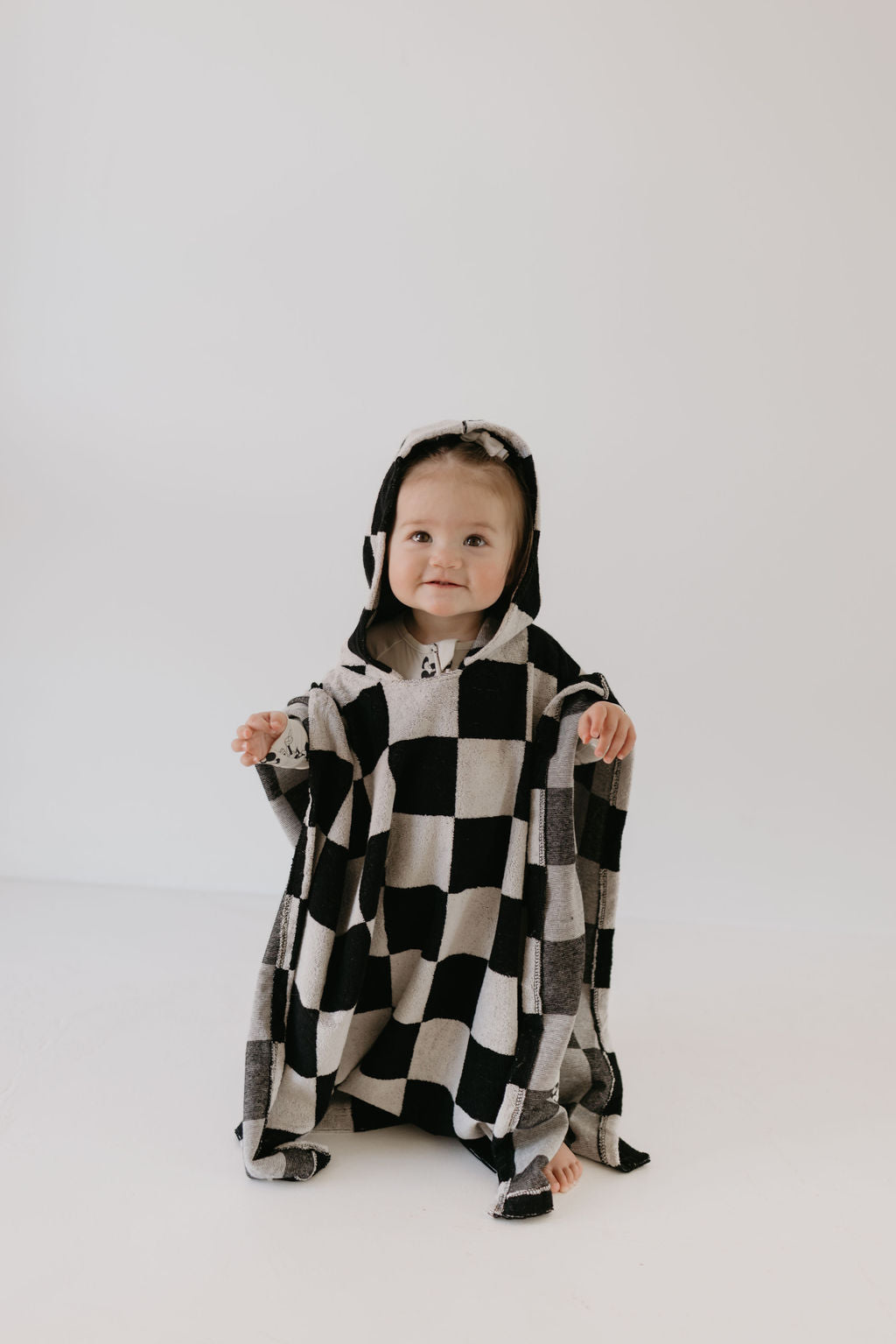 A baby wearing the Hooded Towel in Black Checkerboard from forever french baby sits on a light-colored background, smiling gently. The oversized towel gives a cozy and playful look.