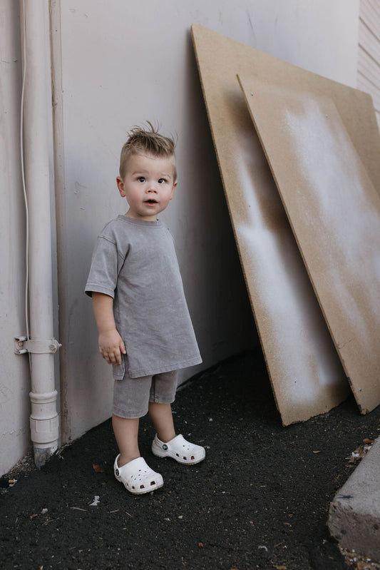 A young child stands outdoors next to a wall with two large boards leaning against it. The child, dressed in a Forever French Baby Children's Short Set in Vintage Washed Steel made from organic cotton, wears a grey t-shirt, matching grey shorts, and white clogs. The child has light brown hair styled upward and looks directly at the camera with a surprised expression.