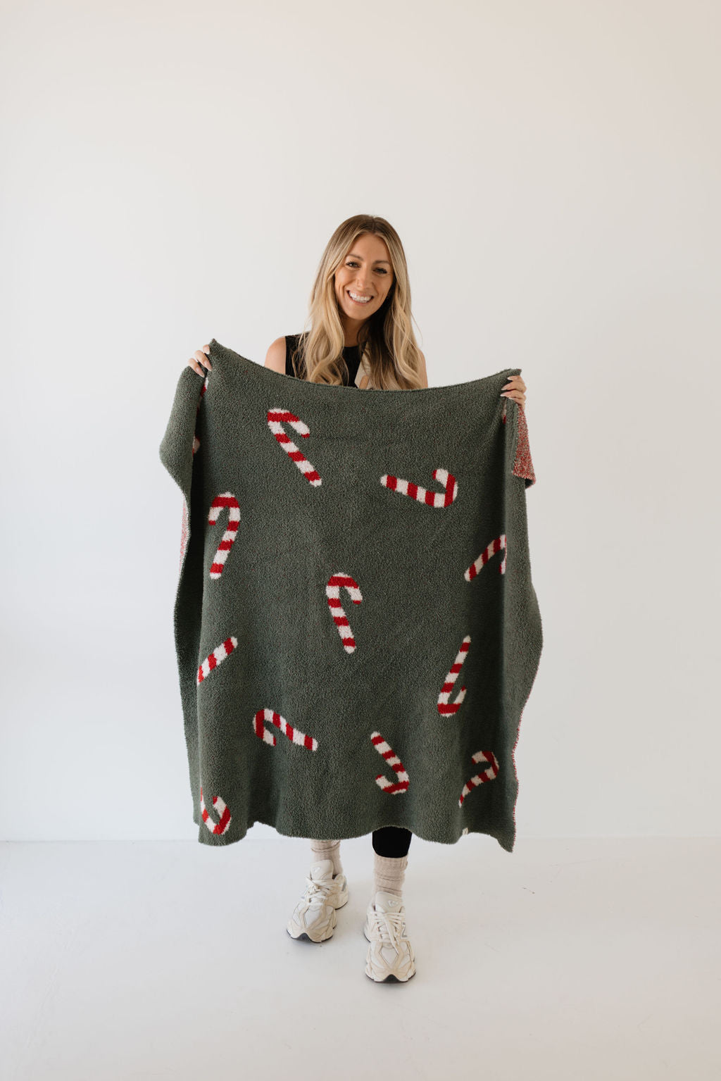 A person stands smiling, holding a Plush Blanket from the forever french baby Candy Cane Lane collection, adorned with red and white candy canes. They're dressed in a black outfit and white sneakers against a plain white backdrop, highlighting their festive spirit.