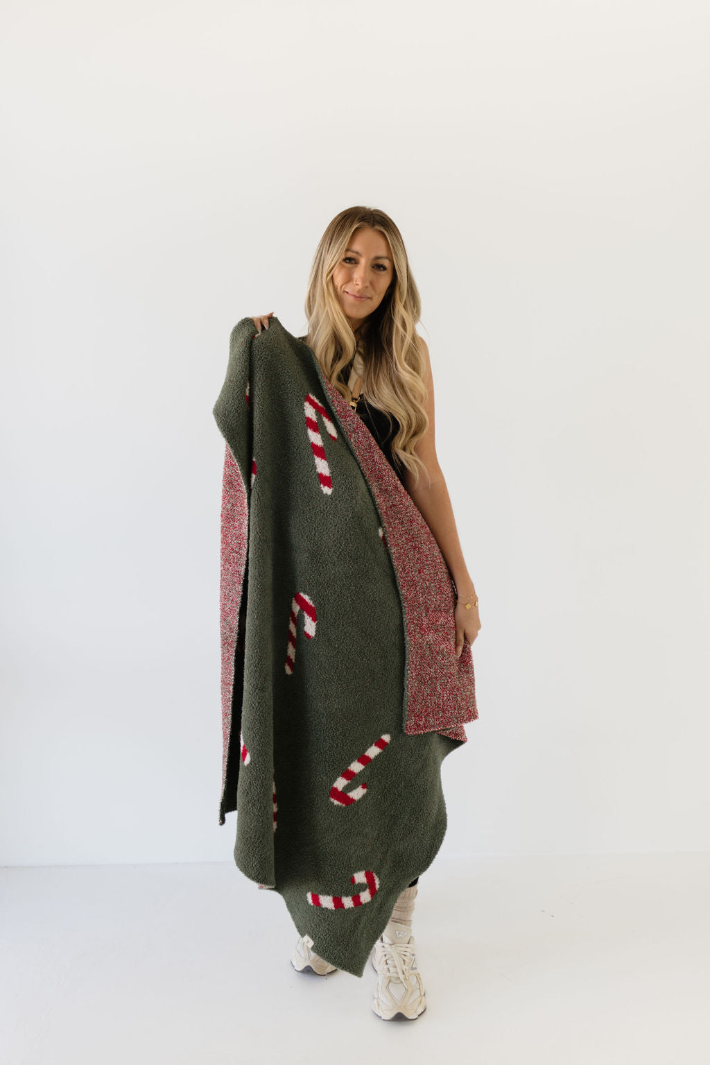 A person with long hair is holding the Plush Blanket from the Candy Cane Lane collection by forever french baby, which features festive red and white candy canes on a green background. They are wearing white sneakers and posing against a plain white background.