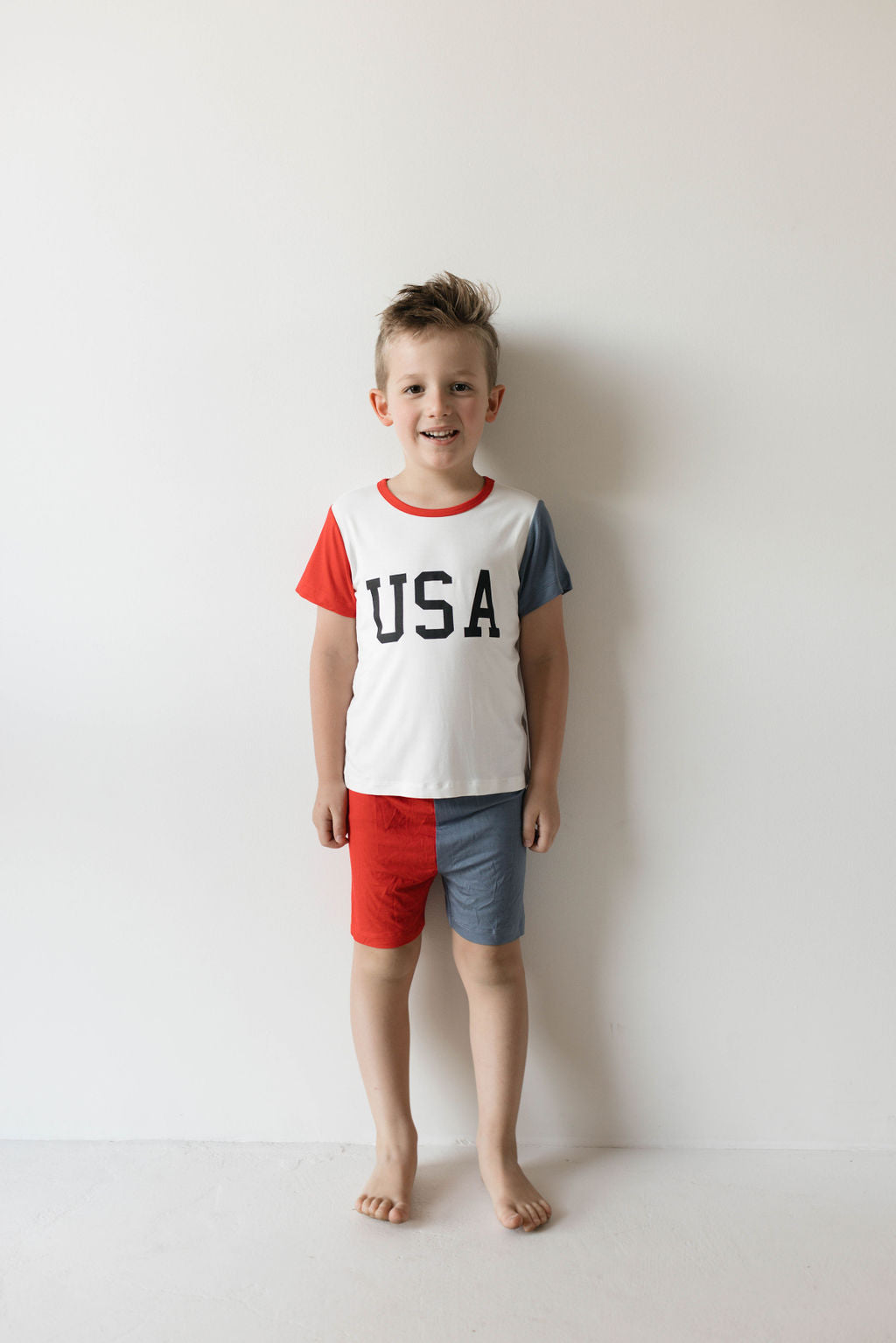 A young boy stands against a plain white background, smiling at the camera. He is wearing a short-sleeved shirt with "USA" printed on it, featuring red and blue sleeves. His mismatched shorts have one red leg and one blue leg. He is barefoot, proudly showcasing his comfy kids pajamas from the forever french baby Bamboo Short Set | USA, Land That I Love collection.