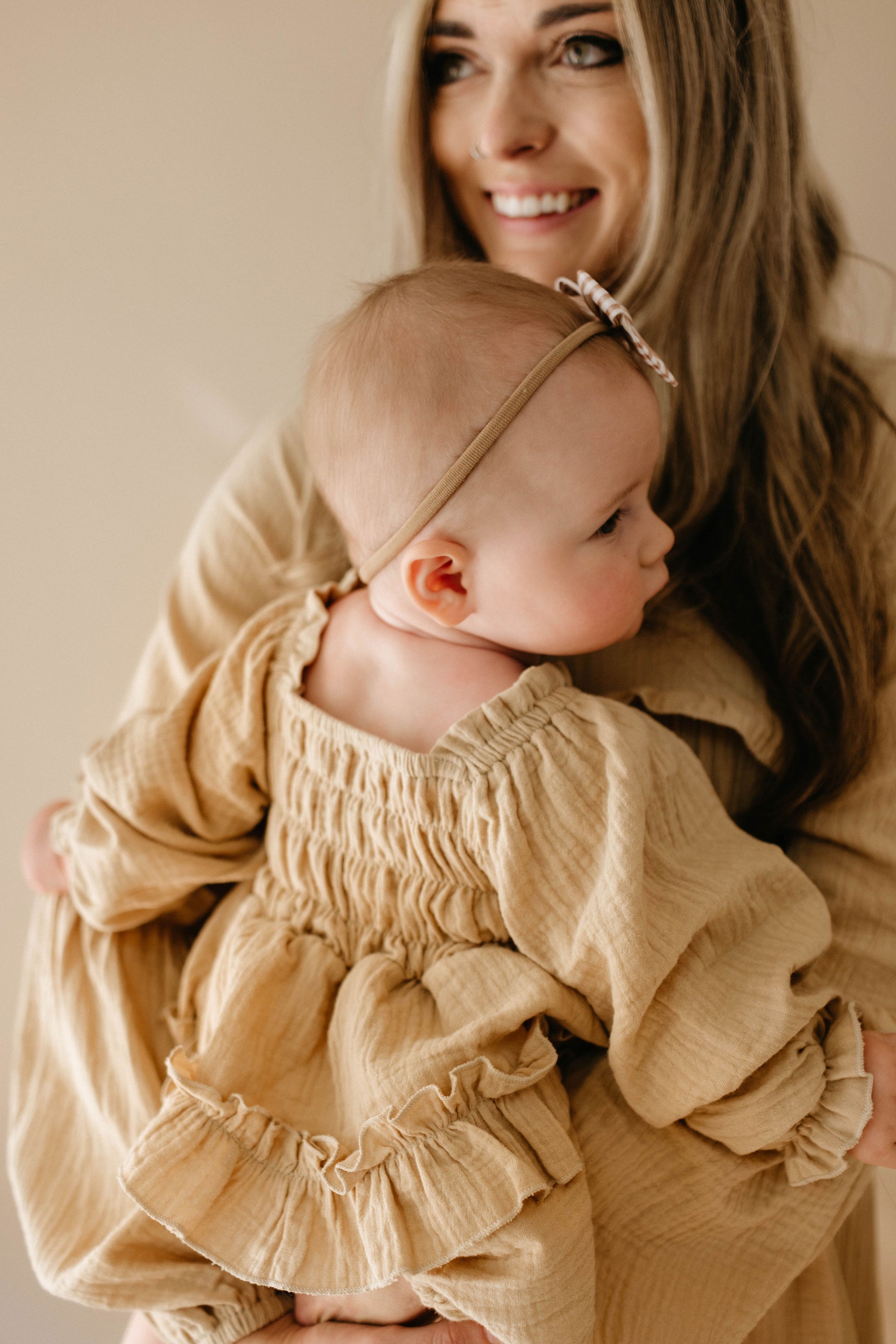 A woman with long hair and a smile holds a baby clad in the "Ruffle Romper | Golden Hour" by forever french baby, complete with a headband. While the baby faces away, their matching neutral-toned outfits harmoniously enhance the golden tones of the softly blurred background.