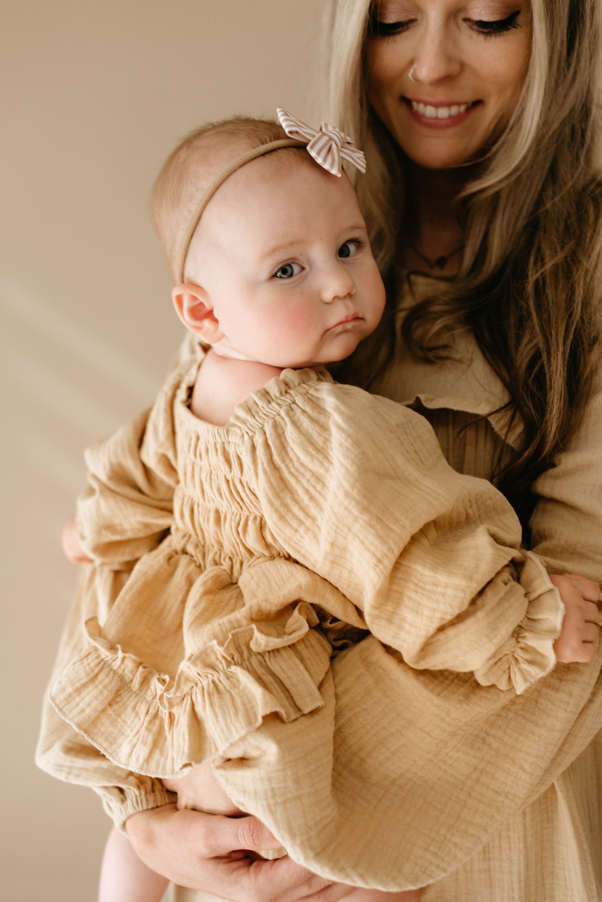 A woman holds a baby in her arms, dressed in the "Golden Hour" Ruffle Romper by forever french baby, complete with a headband featuring a bow. Both smile softly against the neutral beige background, highlighted by a golden hue that gently illuminates their serene moment.