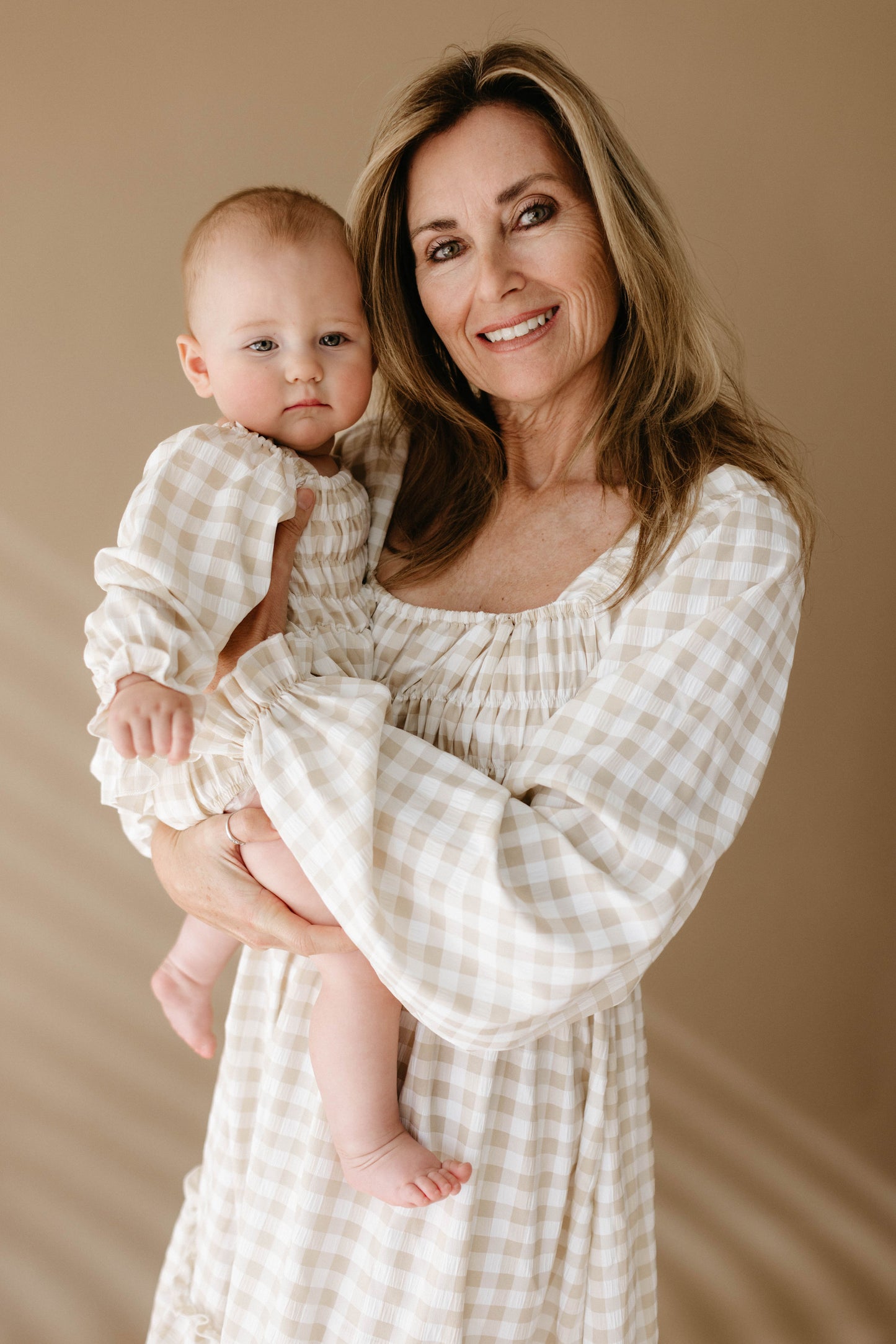 A woman with long hair beams as she holds a baby, both dressed in matching gingham-patterned outfits from forever french baby. The baby's Ruffle Romper and her dress coordinate perfectly against the soft, warm brown backdrop.