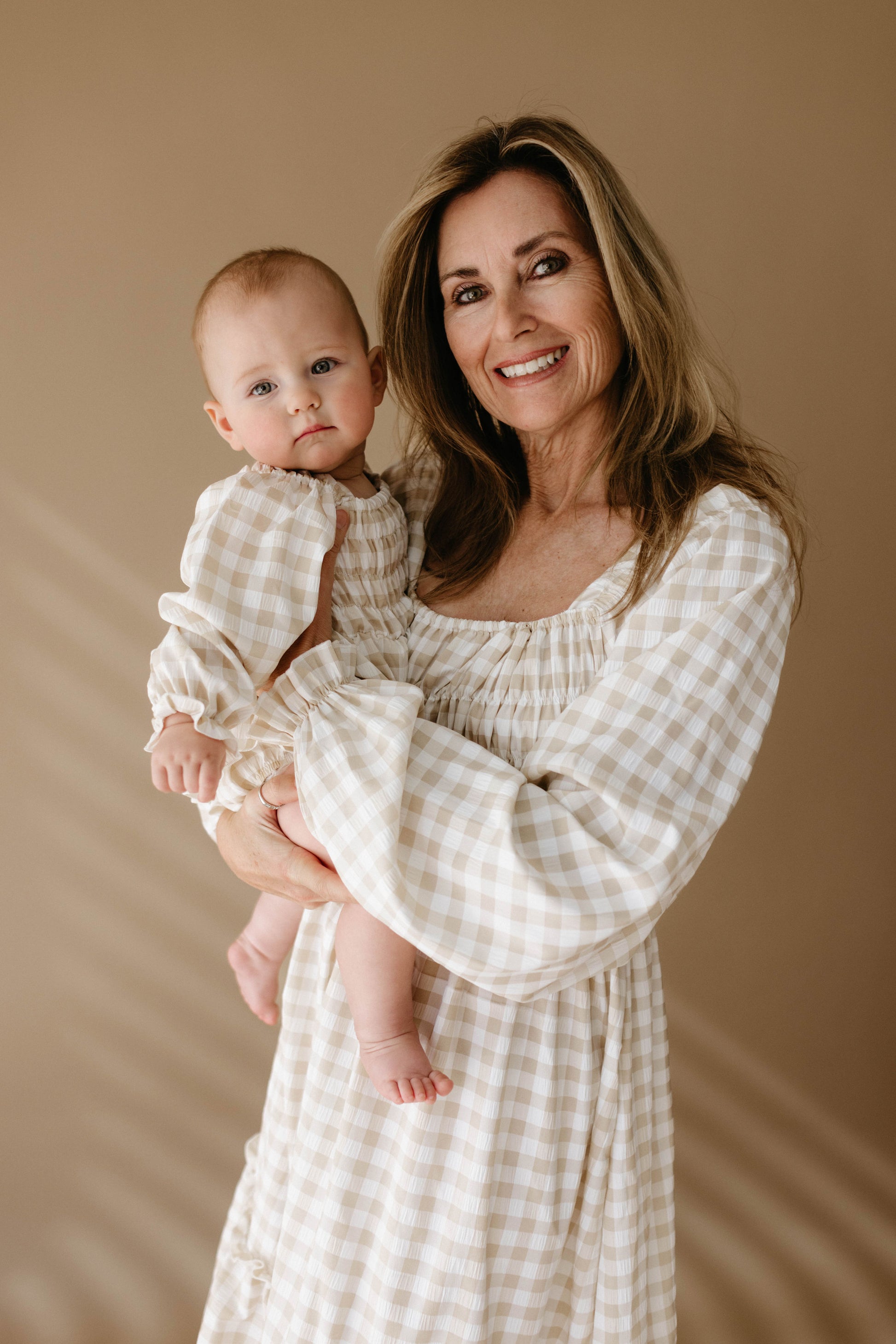 A woman wearing a matching outfit with her baby, both dressed in the forever french baby Ruffle Romper in beige and white gingham, stand against a plain beige background. The woman smiles warmly while the baby gazes at the camera.