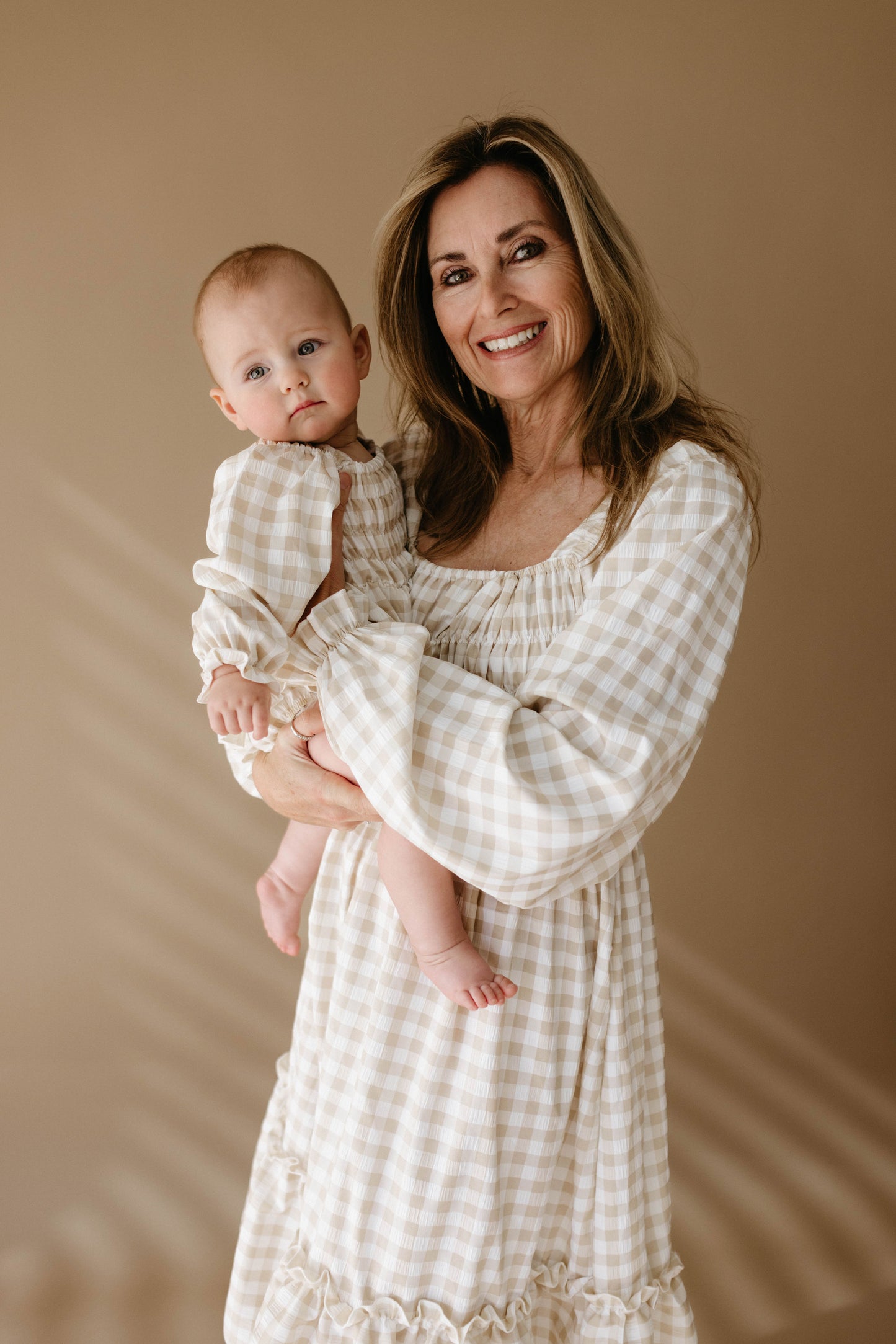 A smiling woman holds a baby in her arms, both dressed in matching beige-toned outfits from the "Ruffle Romper | Gingham" collection by forever french baby. The warm beige background enhances the cozy and harmonious atmosphere.