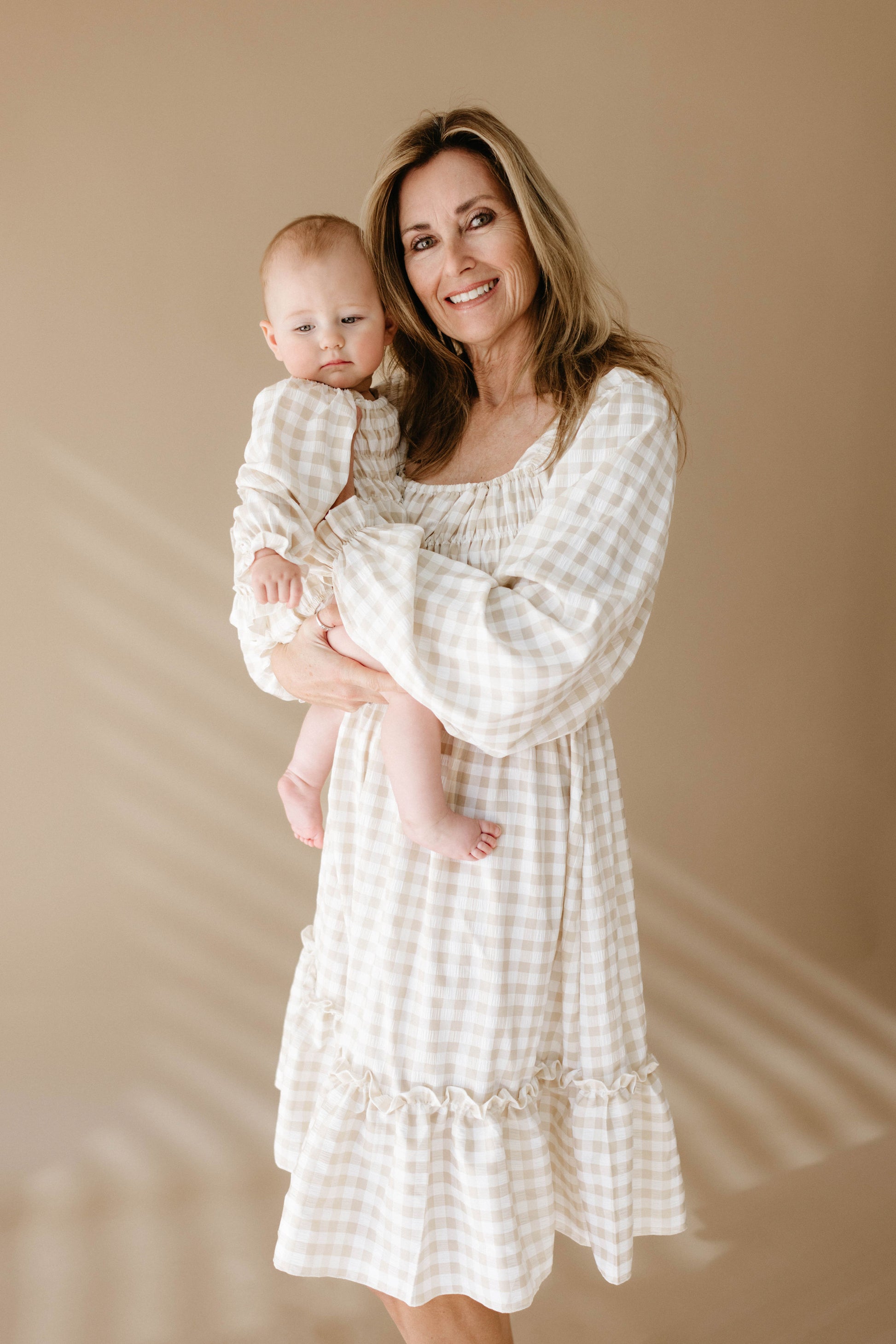 A woman in a light gingham pattern dress smiles while holding a baby dressed in the *forever french baby* Ruffle Romper in Gingham. The background is a plain, warm beige with soft shadows from window blinds gently cast over them.