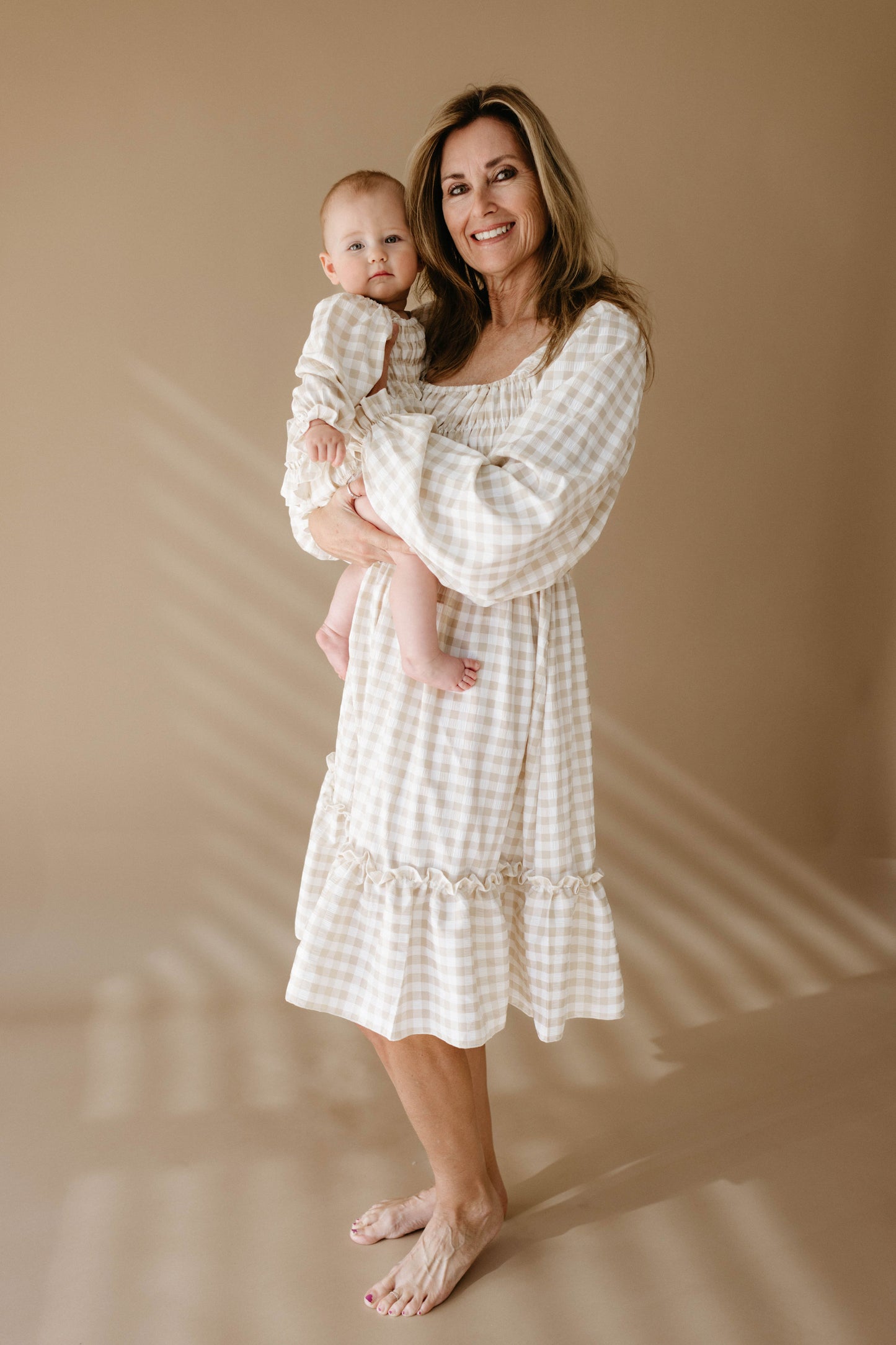 A smiling woman stands barefoot on a light-colored floor, holding a baby. Both are wearing matching outfits from the forever french baby collection, featuring the Ruffle Romper in a beige and white gingham pattern. The background is a plain, warm beige with soft shadows contributing to a cozy atmosphere.