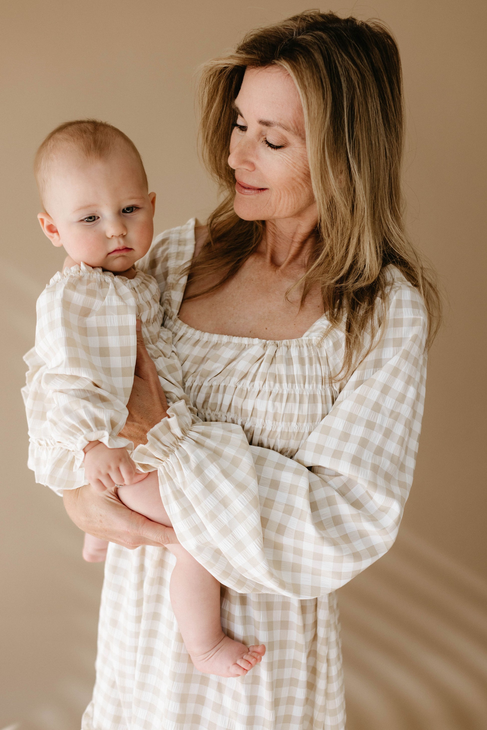 A woman gently holds a baby in her arms, both dressed in matching outfits featuring the Ruffle Romper by forever french baby, showcasing a beige and white gingham pattern. The background is a soft, neutral color, adding warmth to the serene and tender moment.