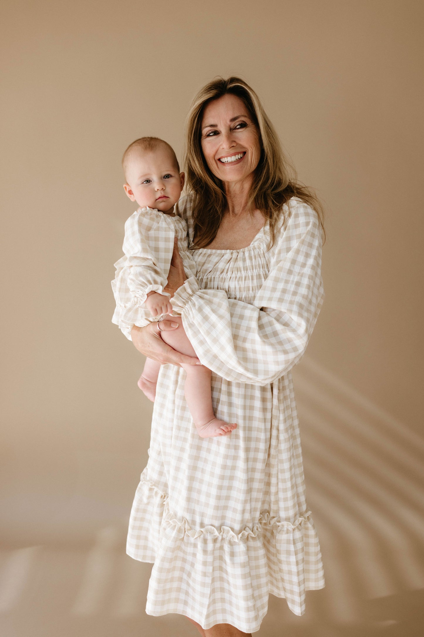 A woman smiles while holding a baby, both dressed in matching Ruffle Romper | Gingham outfits from forever french baby, their neutral tones beautifully complemented by the beige backdrop.