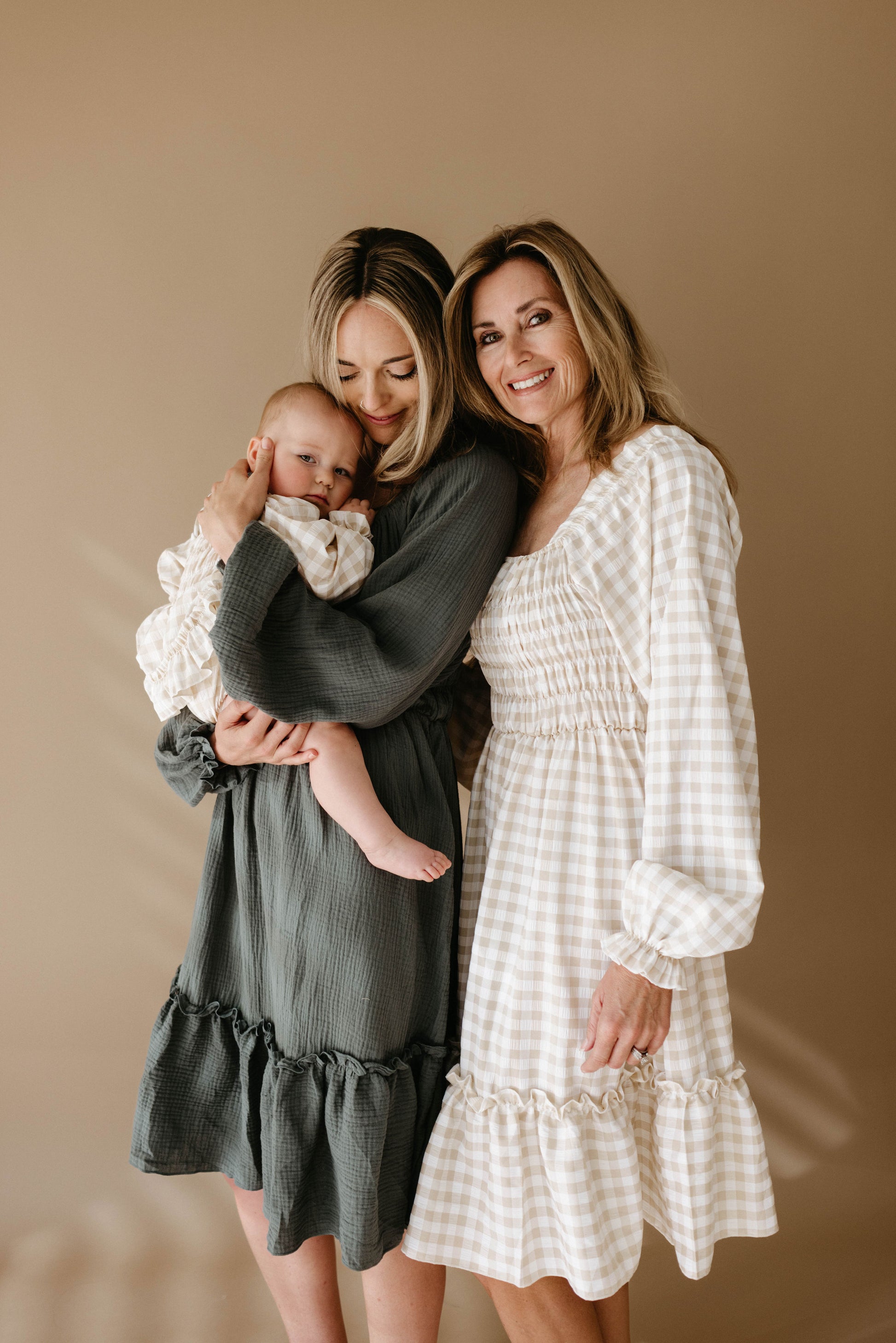 Three generations of women in Meadow long-sleeved ruffled dresses by forever french baby stand together—one holding a baby—all dressed in neutral tones against a simple beige background.