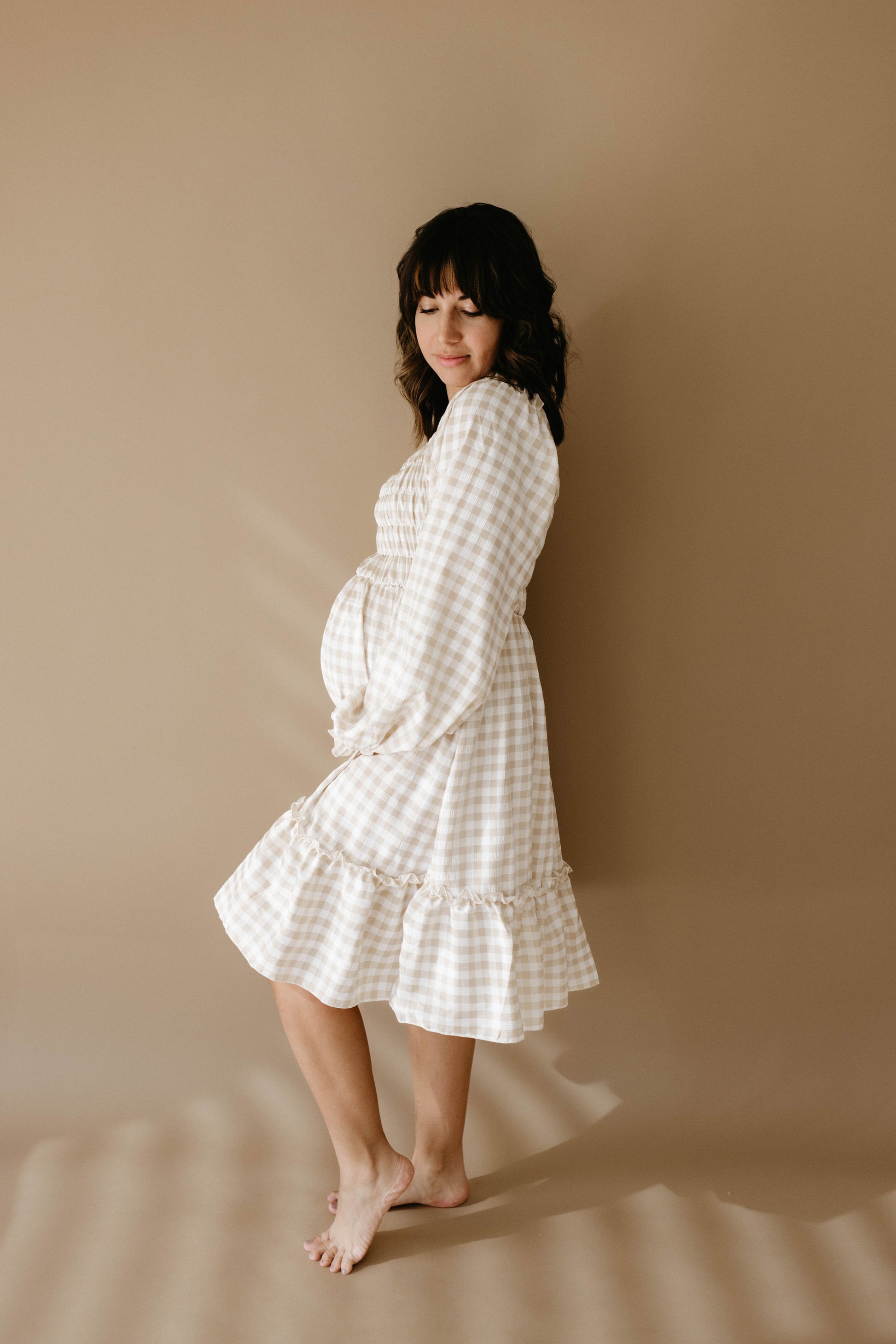 A pregnant woman wearing the Women's Dress in Gingham from forever french baby stands barefoot against a beige backdrop. She gently cradles her belly, looking downward with a soft smile. Nearby, matching rompers and child-size dresses suggest the joyous future to come. Her dark hair cascades in loose waves.