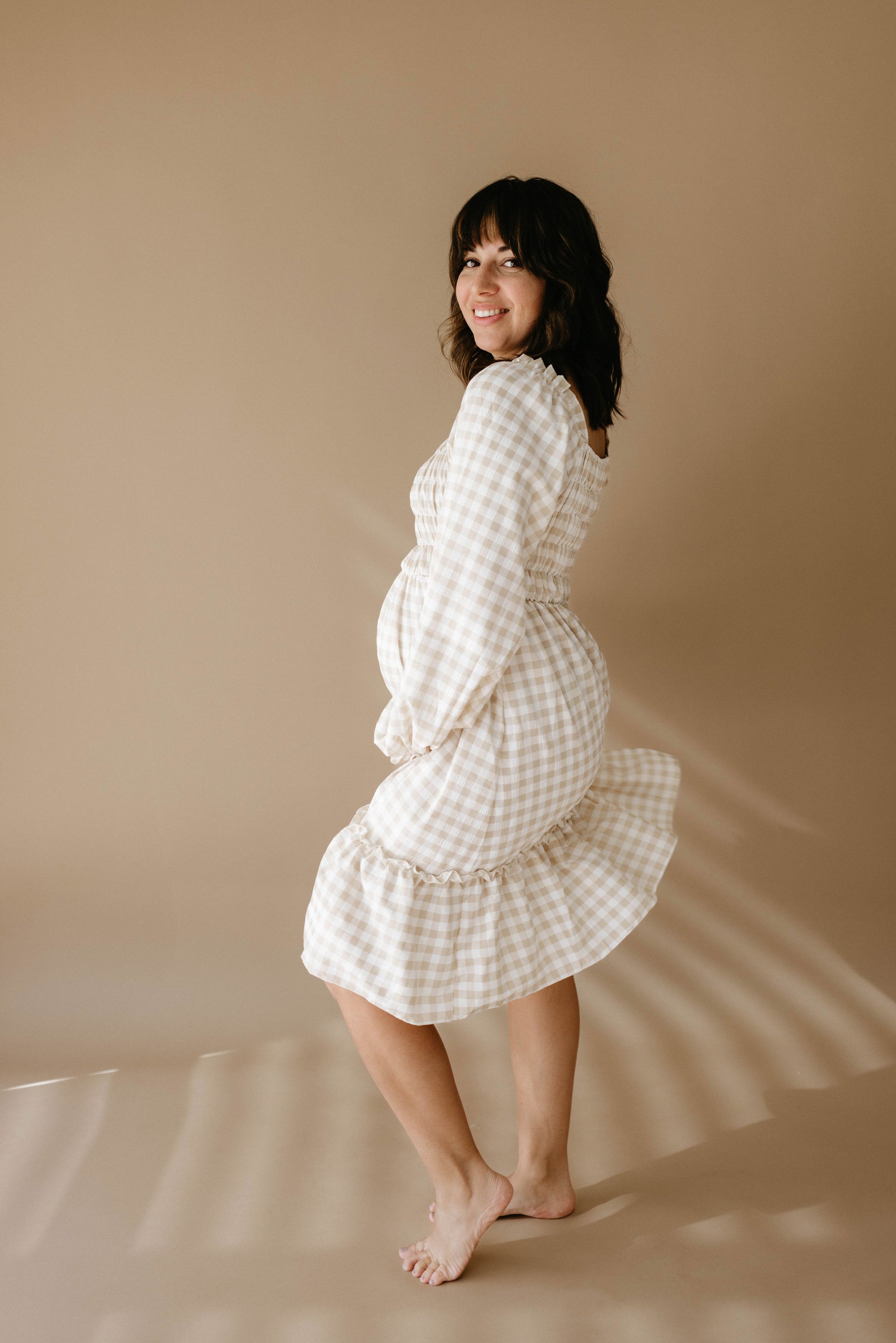 A woman stands barefoot on a beige background, wearing the Women's Dress | Gingham by forever french baby. She is smiling with one hand on her hip and the other gently touching her belly, hinting at her pregnancy. Soft light casts subtle shadows around her.