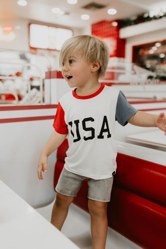 A young child with blonde hair, wearing a vibrant Children's Bamboo T-Shirt from forever french baby that says "USA, Land That I Love" and shorts, stands in a brightly lit diner with red and white decor. The child is smiling with an enthusiastic expression, embodying the spirit of 4th of July clothes in an eco-friendly shirt.