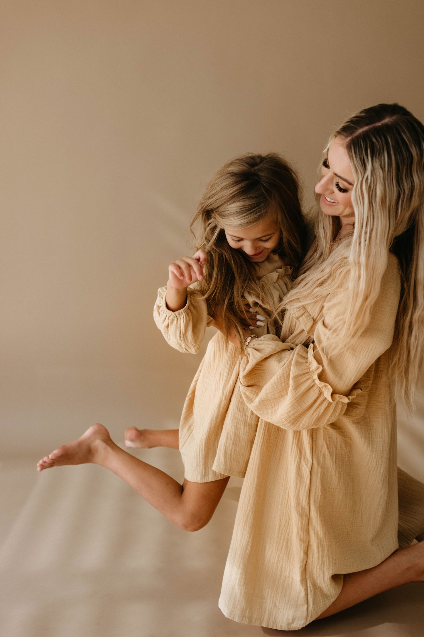 A woman and a young girl, both wearing matching "Golden Hour" outfits from forever french baby, are laughing and hugging against a neutral background. The woman has long hair, and the girl looks joyful as she leans into the embrace, their child-size dresses adding to the heartwarming scene.